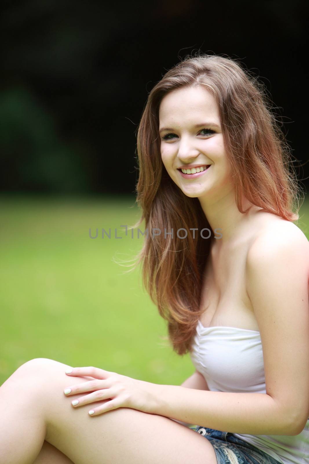 Portrait of a pretty teenage girl sitting on the grass in a lush park smiling at the camera