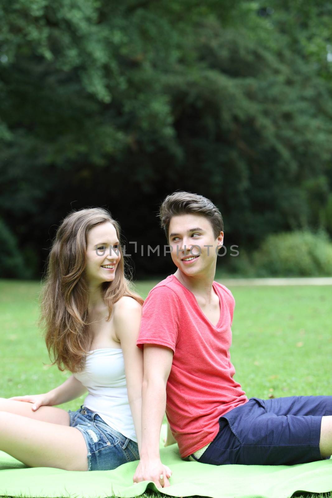 Young teenage couple in the park sitting back to back on the grass looking over their shoulders at each other in a lush green park