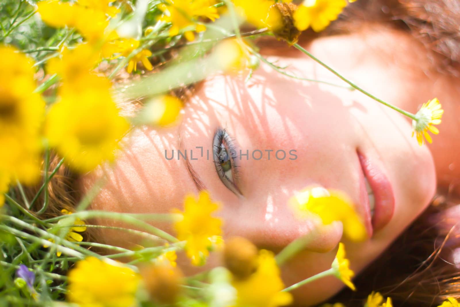 Beautiful girl laying on a field among fresh grass & yellow flowers.