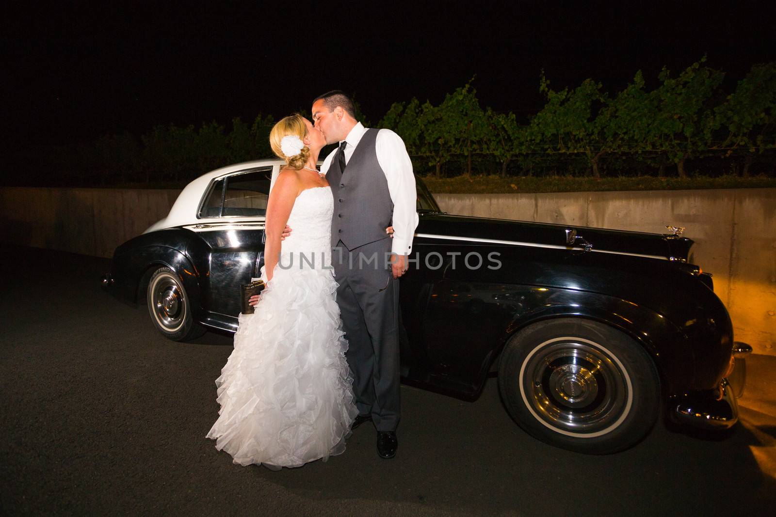 A bride and groom pose for a last photo in front of an old classic car at a vineyard at night.