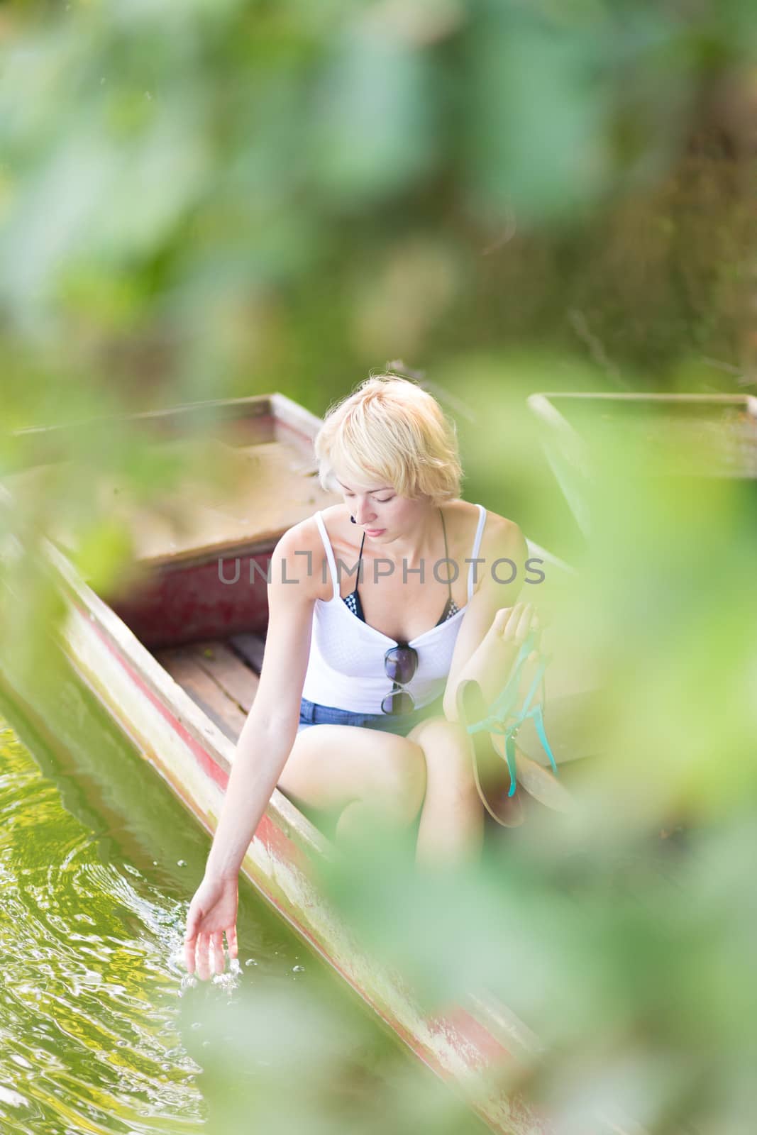 Thoughtful young blonde woman enjoying the sunny summer day on a vintage wooden boats on a lake in pure natural environment on the countryside.