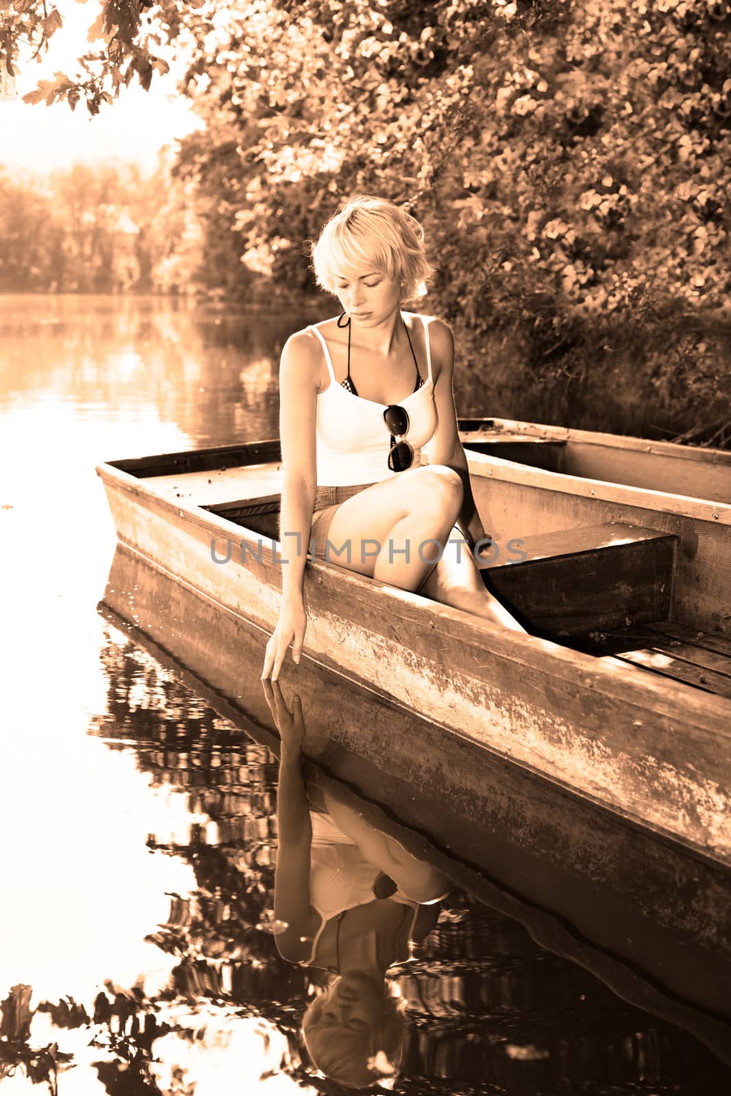 Thoughtful young blonde woman enjoying the sunny summer day on a vintage wooden boats on a lake in pure natural environment on the countryside. Sepia toned image.