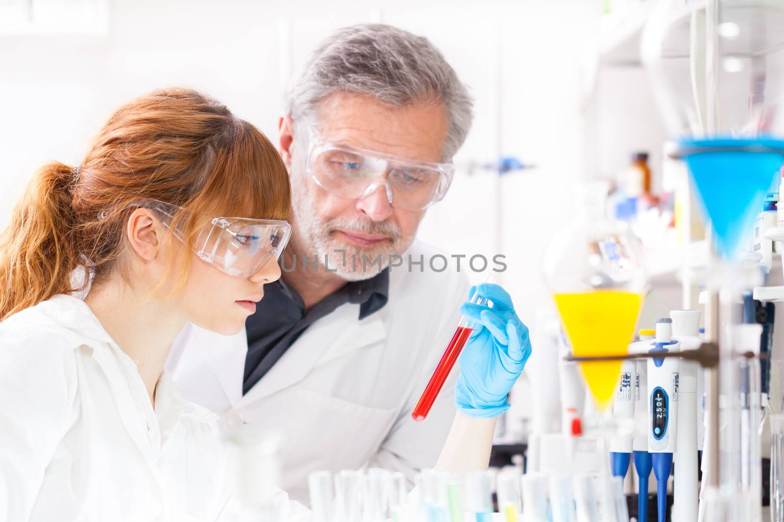 Attractive young female scientist and her senior male supervisor observing color shift of a red liquid in the glass tube in the life science research laboratory (bichemistry, genetics, forensics, microbiology..)