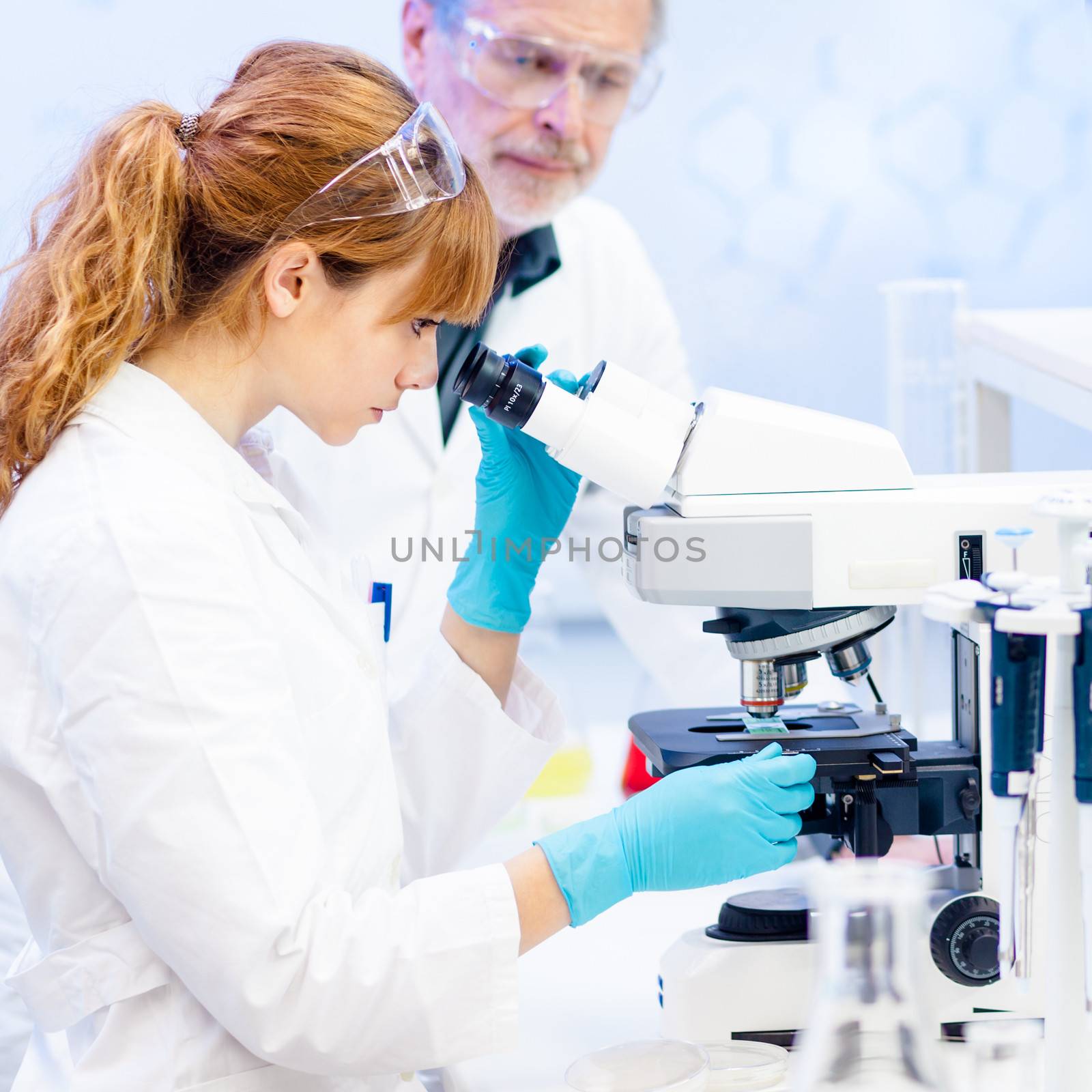 Attractive young female scientist and her senior male supervisor looking at the microscope slide in the life science research laboratory (biochemistry, genetics, forensics, microbiology..)