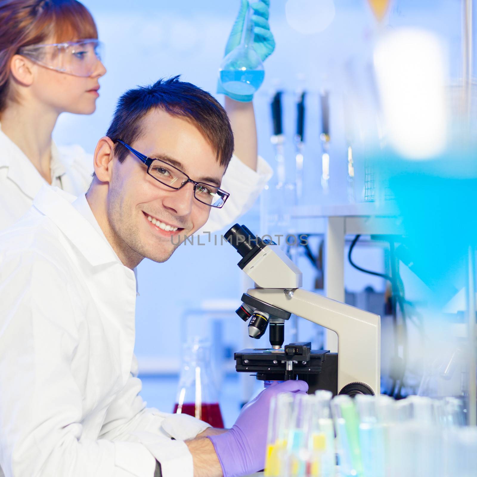 Portrait of a young male researcher microscoping in the life science (forensics, microbiology, biochemistry, genetics, oncology...)laboratory. Female asistant scientist examining blue liquid solution at the beaker in the background.