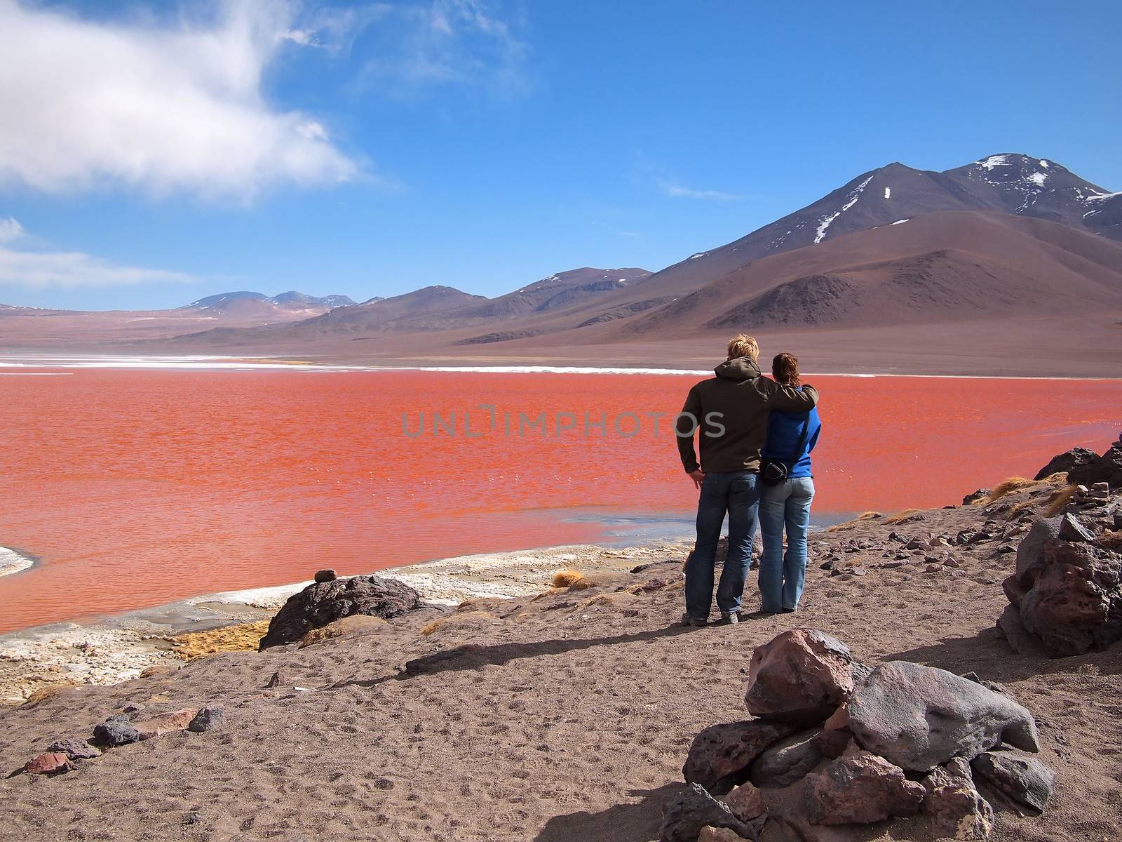 Young tourist couple admiring the Red Lagoon, or Laguna Colorada, on the Altiplano near Uyuni inside Eduardo Avaroa National Reserve in Bolivia at 4300 m above sea level.  The red color of the water is caused by sediments and algae. The white part is borax salt.