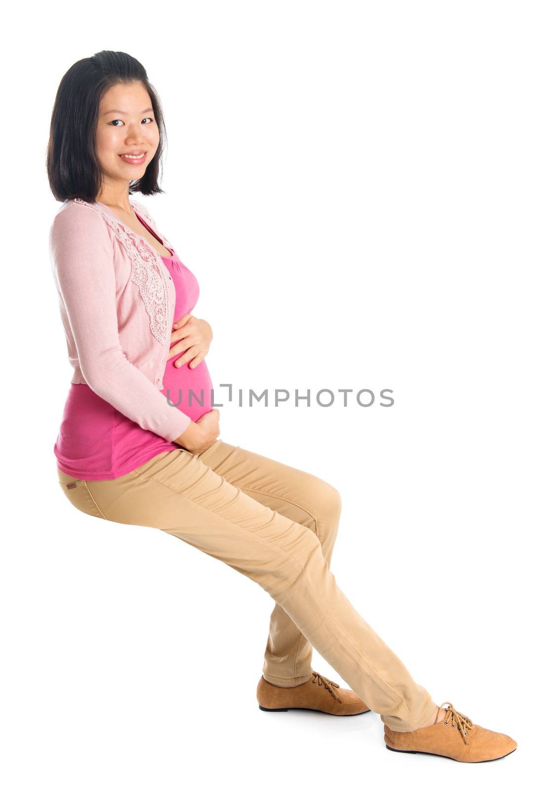 Full body six months pregnant Asian woman sitting on invisible chair, looking at camera,  isolated on white background.