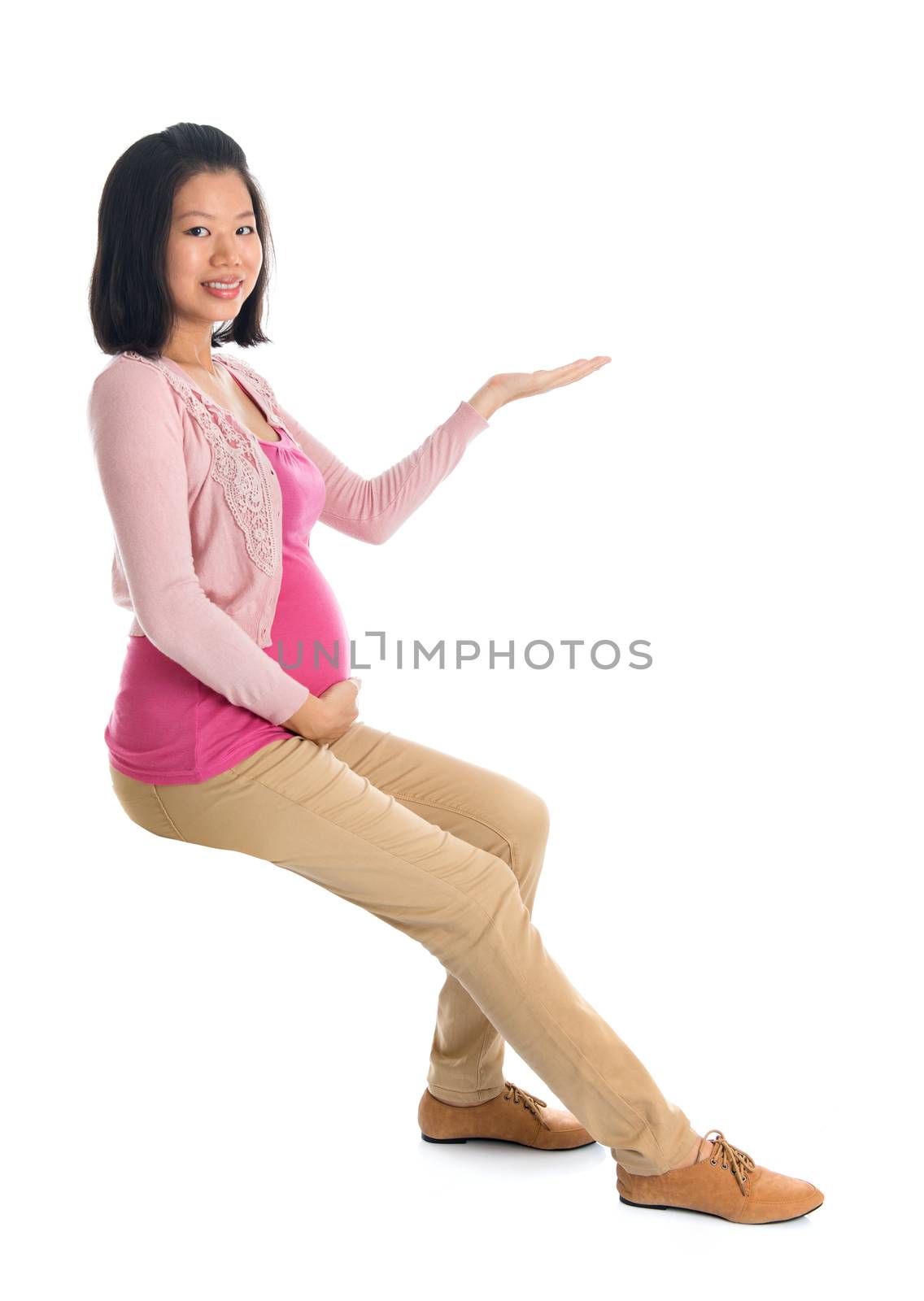 Full body six months pregnant Asian woman sitting on invisible chair, hand showing blank space looking at camera,  isolated on white background.