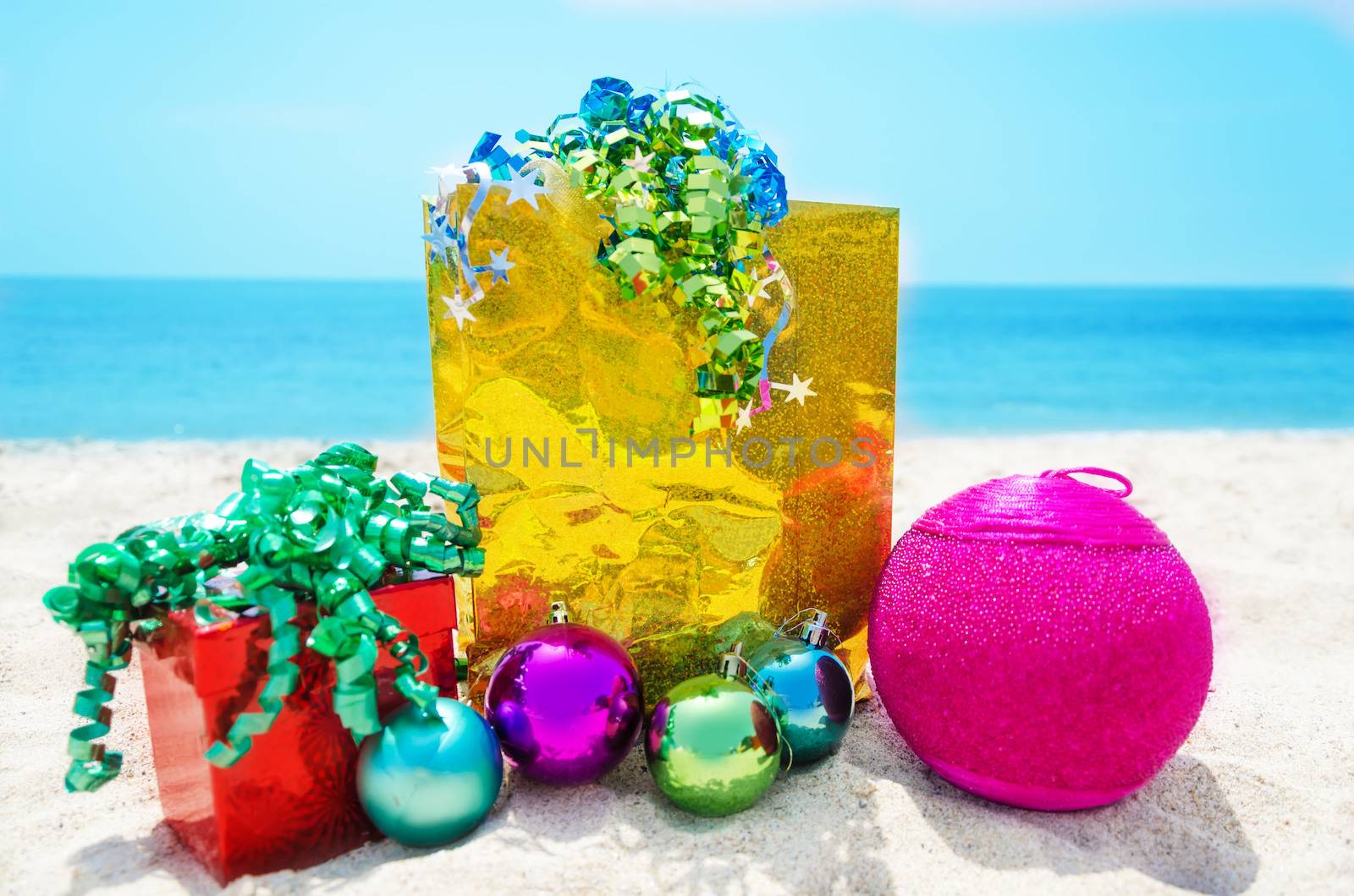 Gold gift bag and red gift box with Christmas balls on sandy beach in sunny day- holiday concept