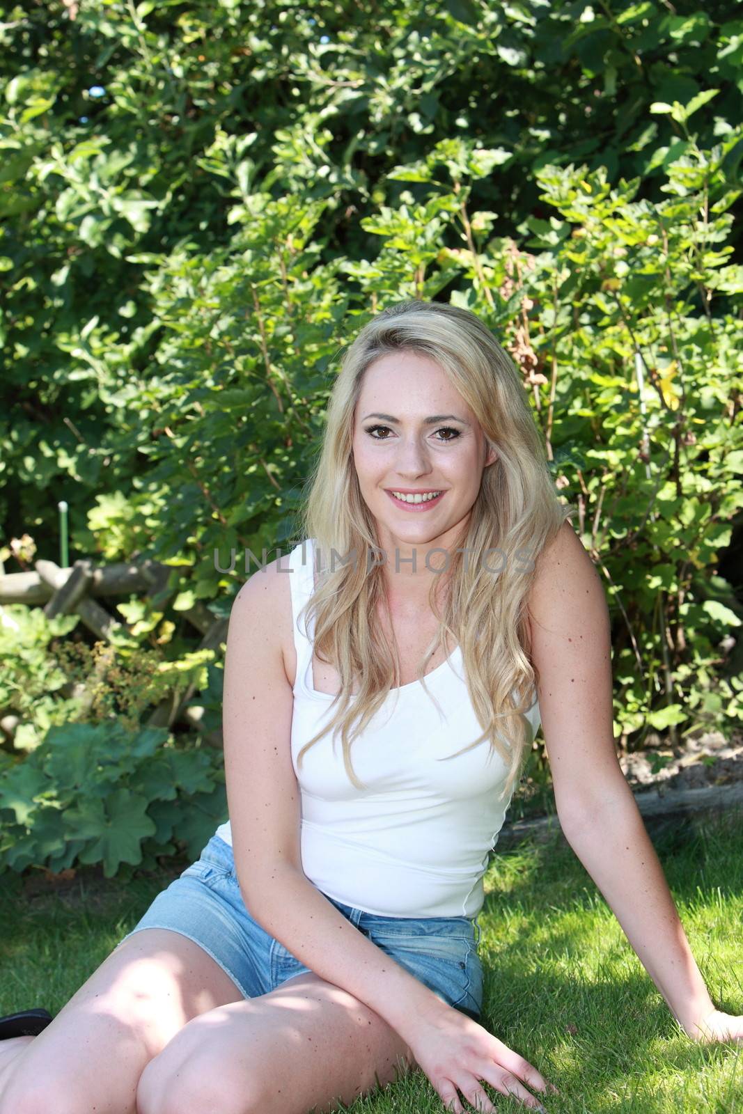 Smiling beautiful woman in the shade of a tree relaxing in denim shorts on the green grass looking at the camera with a smile