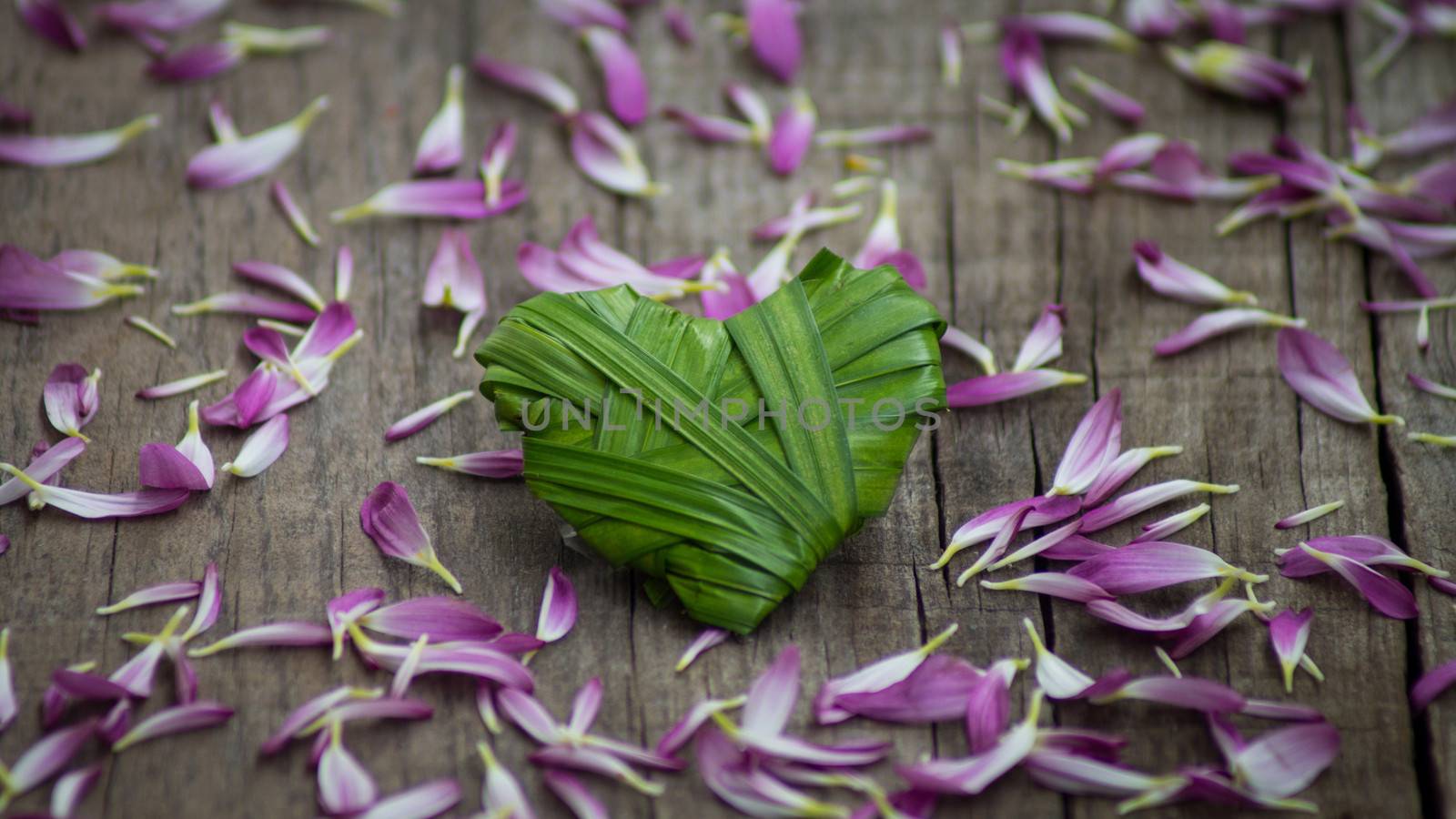 A plam leaf heart surrounded by purple petals on wood background.