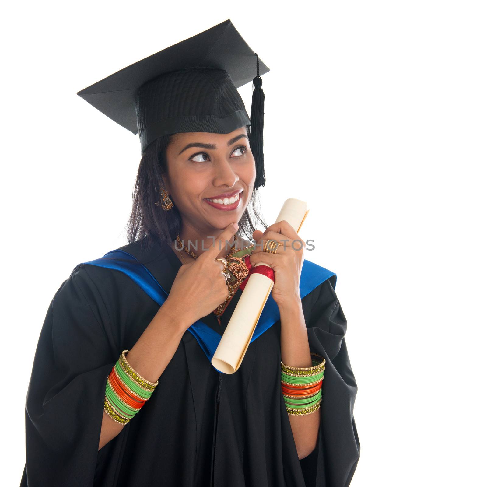 Happy Indian university student in graduation gown and cap holding diploma certificate thinking. Portrait of mixed race Asian Indian and African American female model standing isolated on white background.