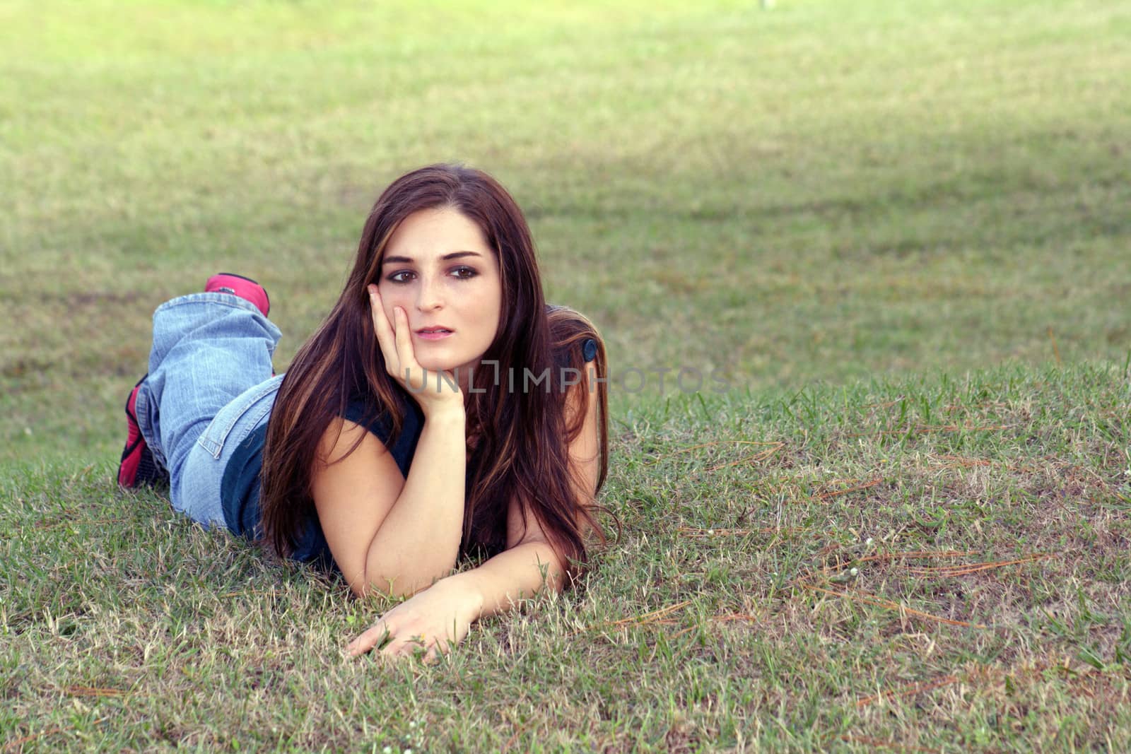 An attractive young brunette in casual wear relaxes outdoors, lying in the grass.