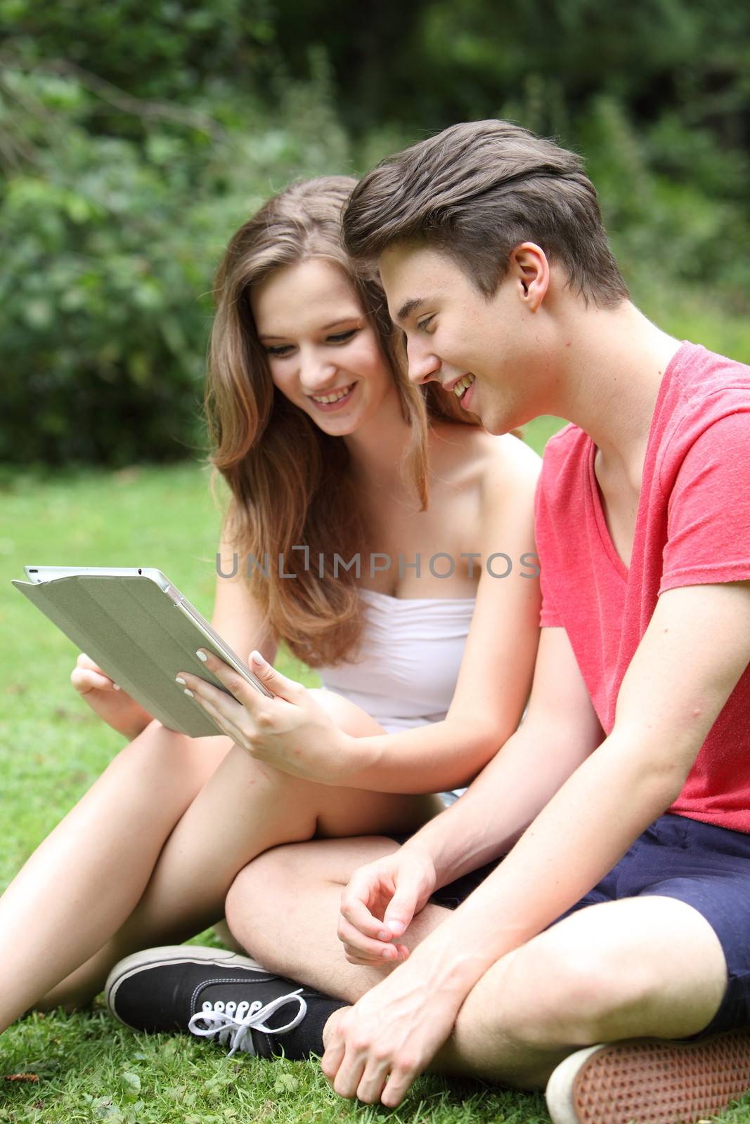 Young teenage girl and boy sitting together on the green grass sharing a tablet-pc as they smile at information on the screen