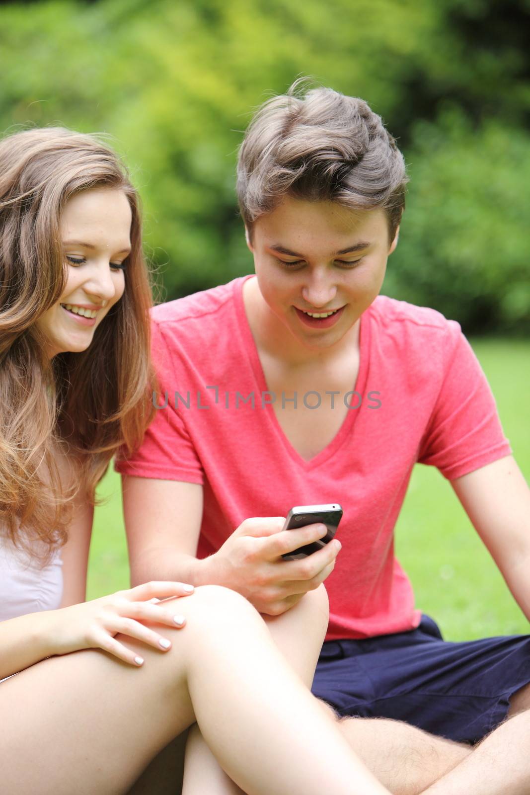 Happy young teenage boy and girl out on a date sitting in a lush green parking reading text messages on their mobile phone