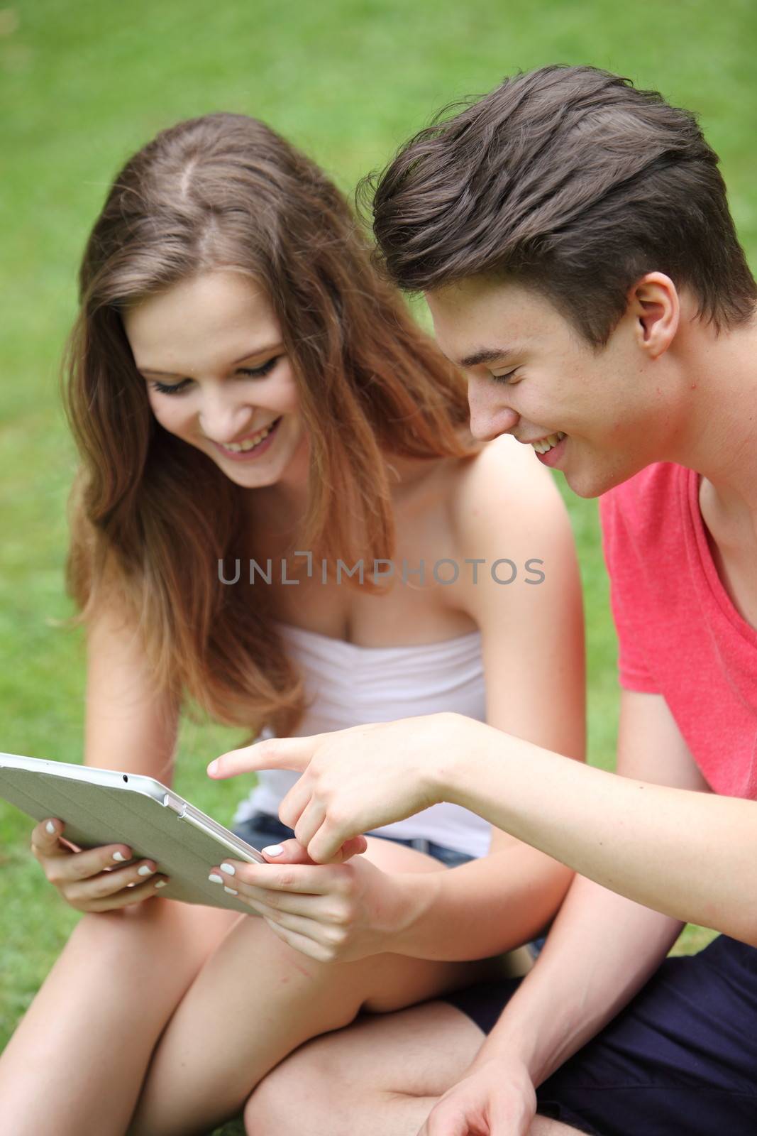Attractive relaxed happy young teenage boy and girl sitting on green grass using a tablet-pc