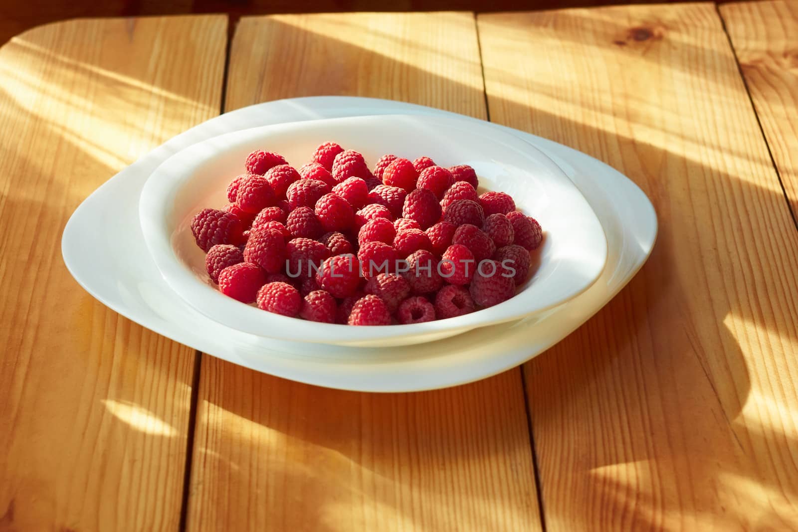 Ripe red raspberries in a plate on the wooden table partially lighted by sunlight