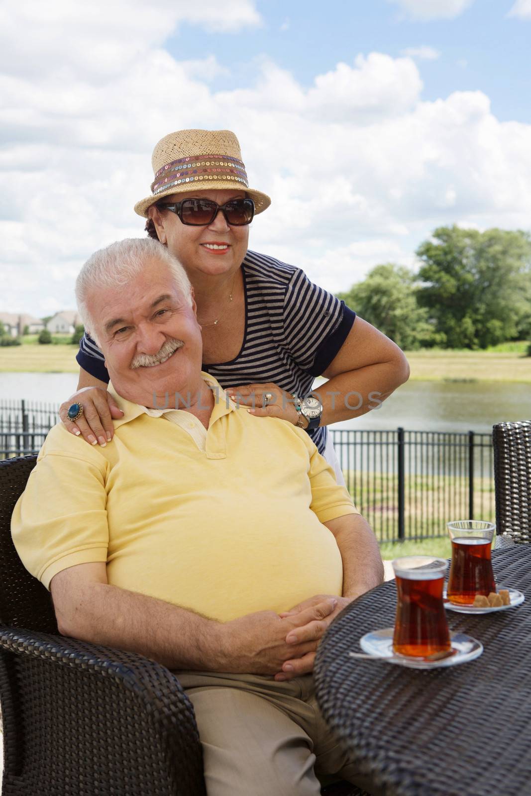 Loving senior couple having drinks on an outdoor patio giving the camera beautiful beaming smiles with the woman posing behind her husband with her hands on his shoulders