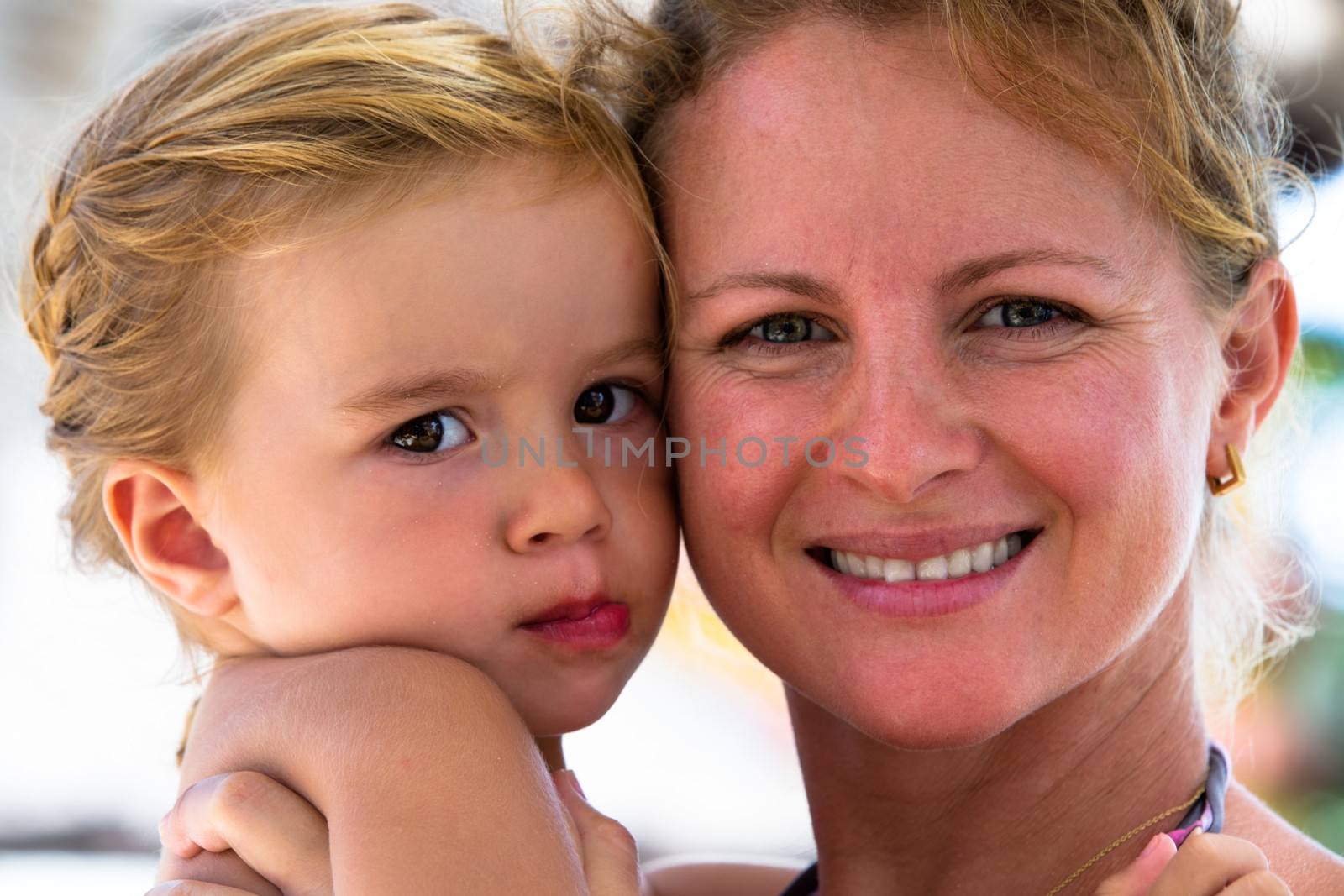 Mother and daughter posing to camera cheek to cheek, Summer time, some sand on their faces.