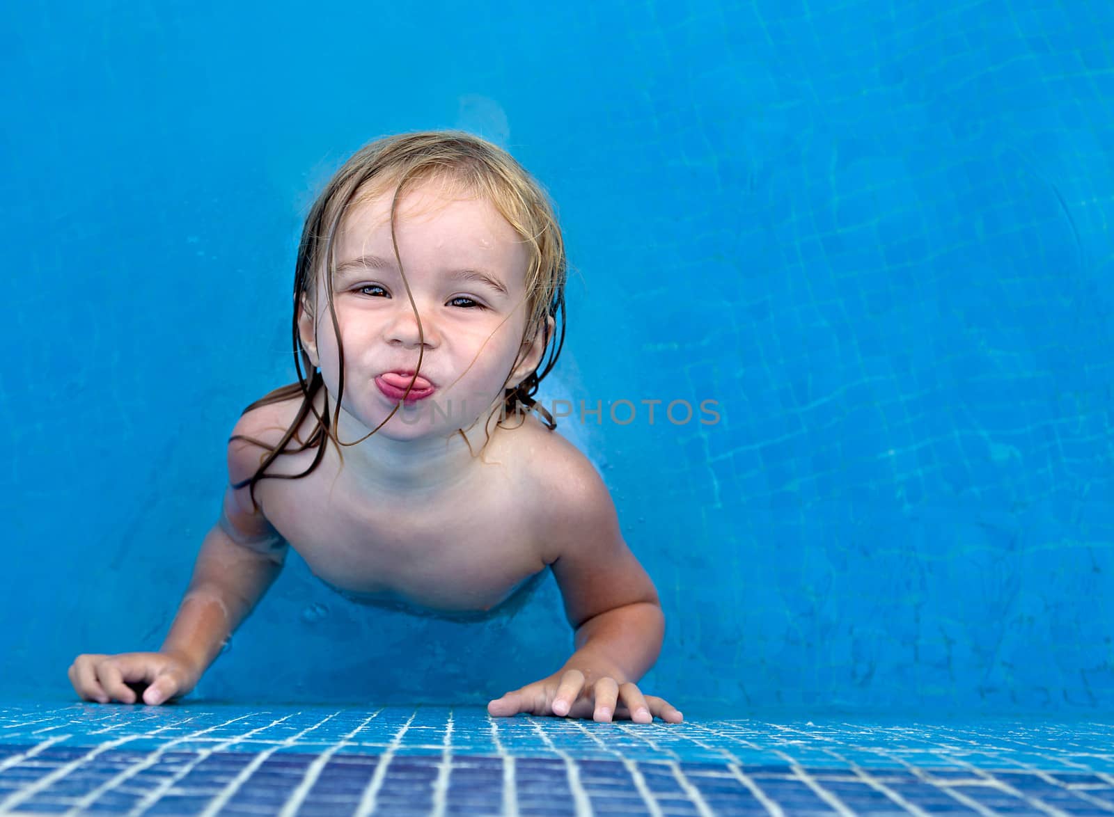 Little toddler girl posing playfully her tongue out from the blue swimming pool, copy space on the right