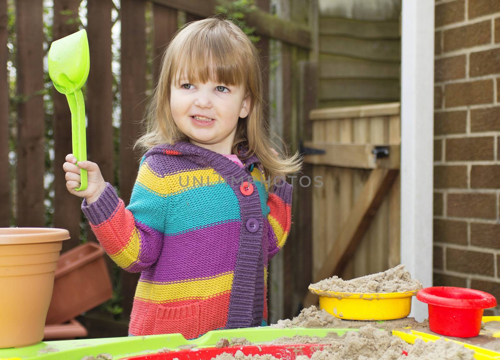 Little girl playing at a sand table in a garden, holding a spade and smiling