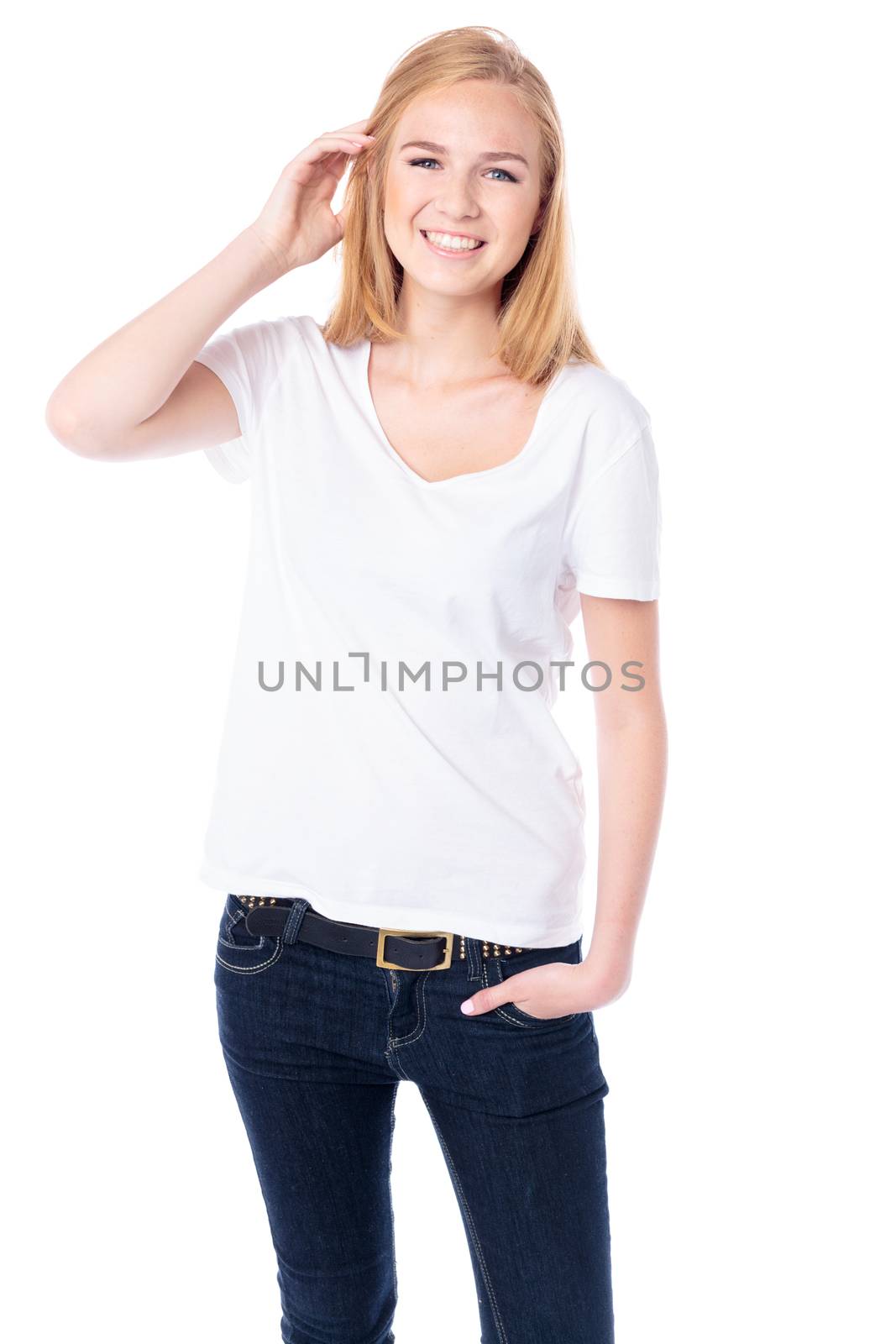 Happy young blond female student in casual jeans standing facing the camera with her hand raised to her hair smiling at the camera, threequarter body on white