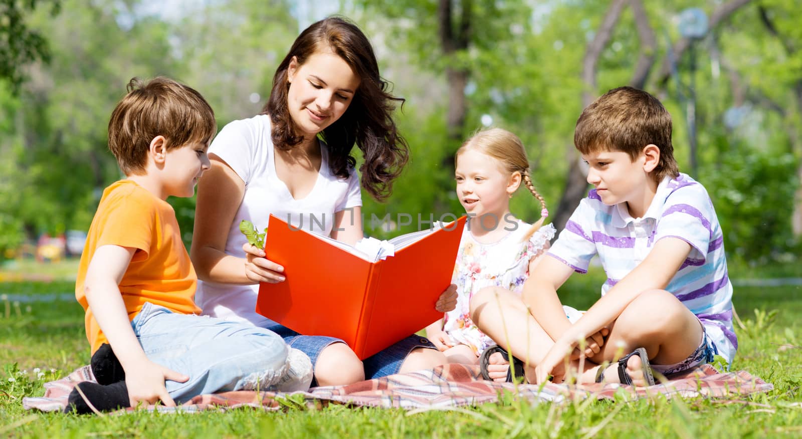 children and teacher reading book together in the summer park