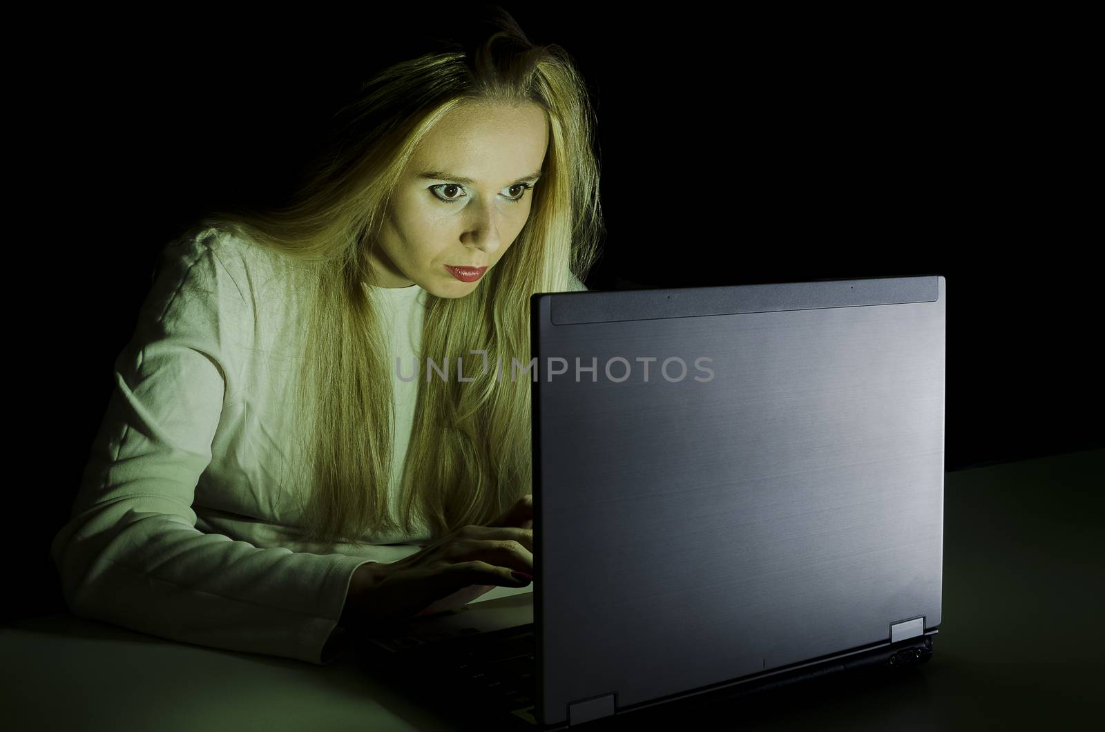 woman working on a computer by night in a dark room with only light from computer falling on her face horizontally cropped