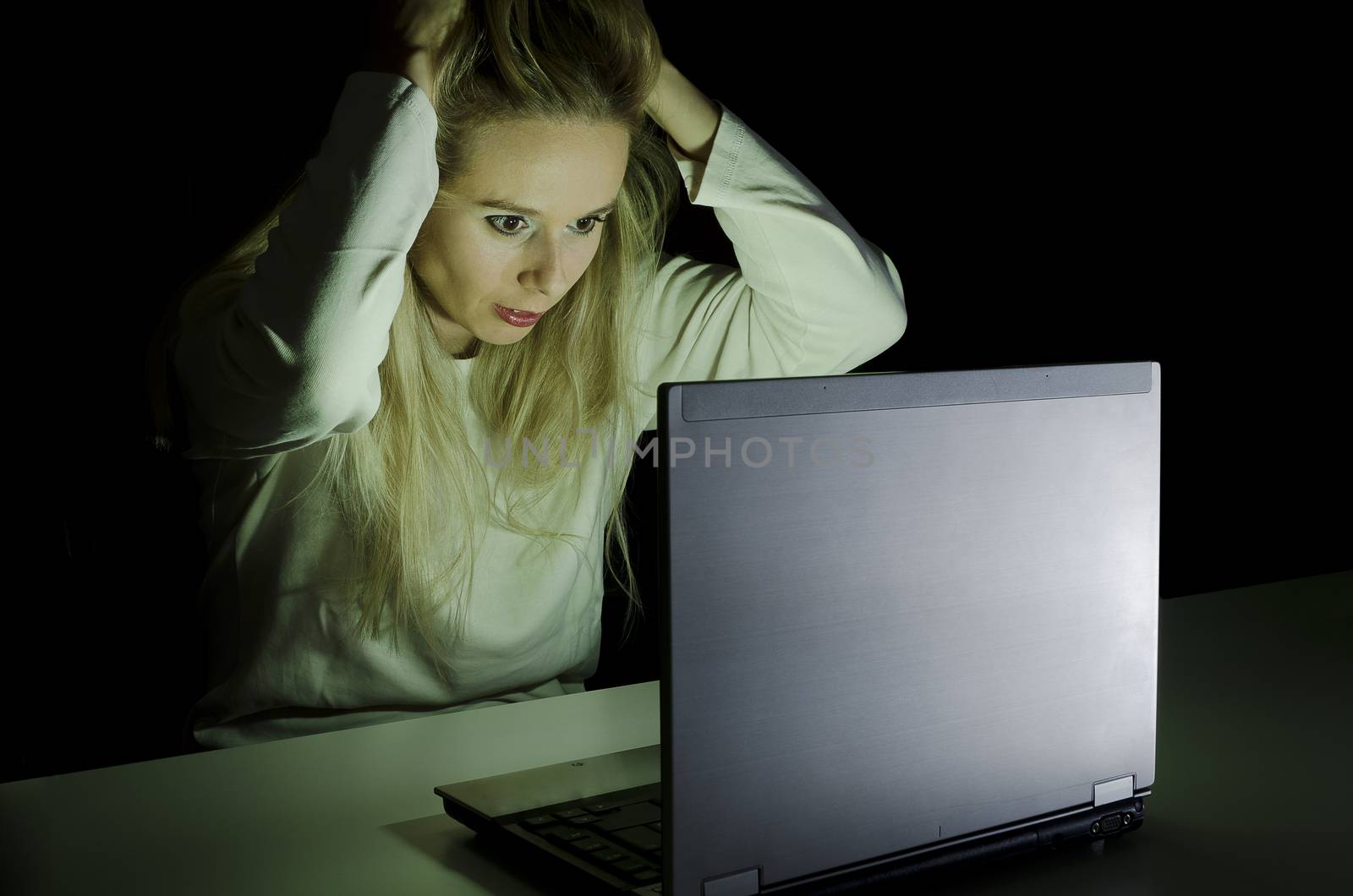 woman working on a computer by night in a dark room with only light from computer falling on her face pulling her hair in frustration