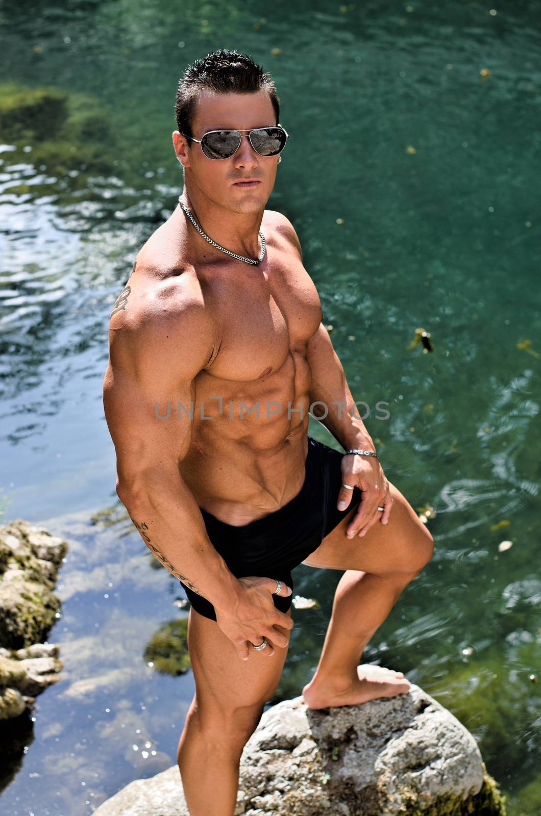 Handsome young muscle man standing in water pond shirtless with sunglasses