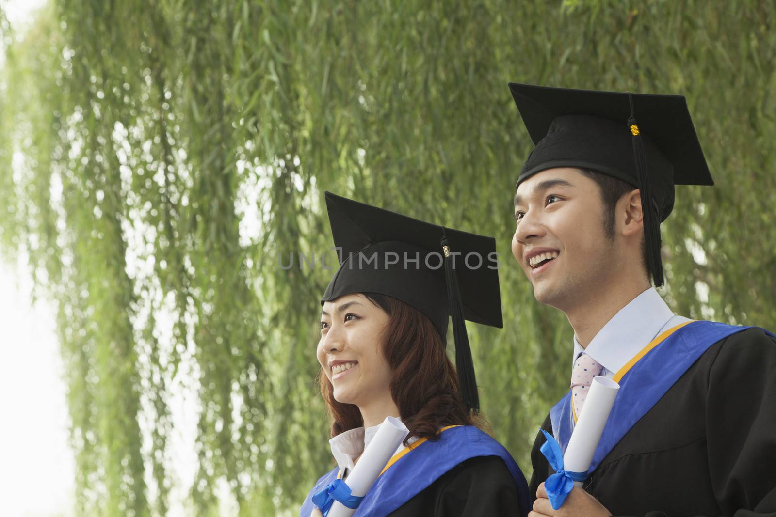 Two Young University Graduates Holding Diplomas And Looking Away 