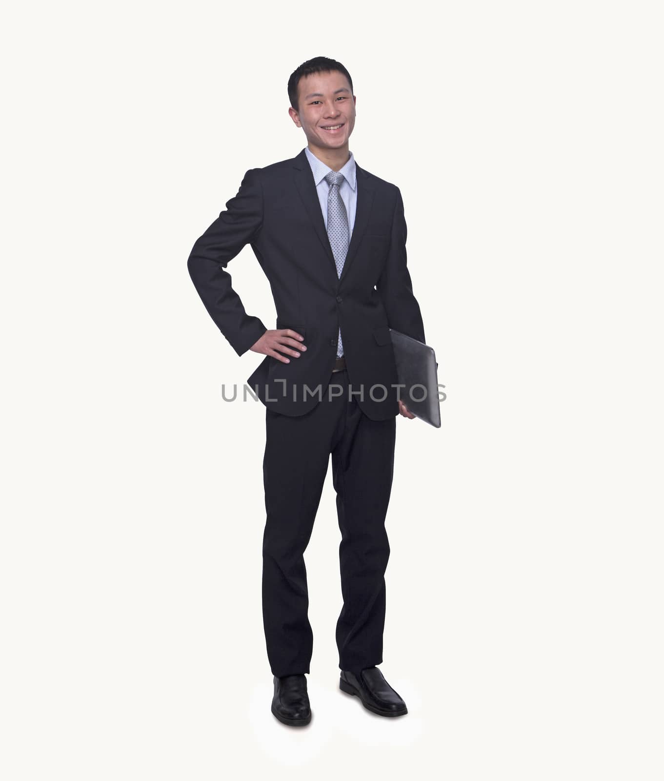 Portrait of smiling young businessman holding a laptop, hand on hip, studio shot by XiXinXing