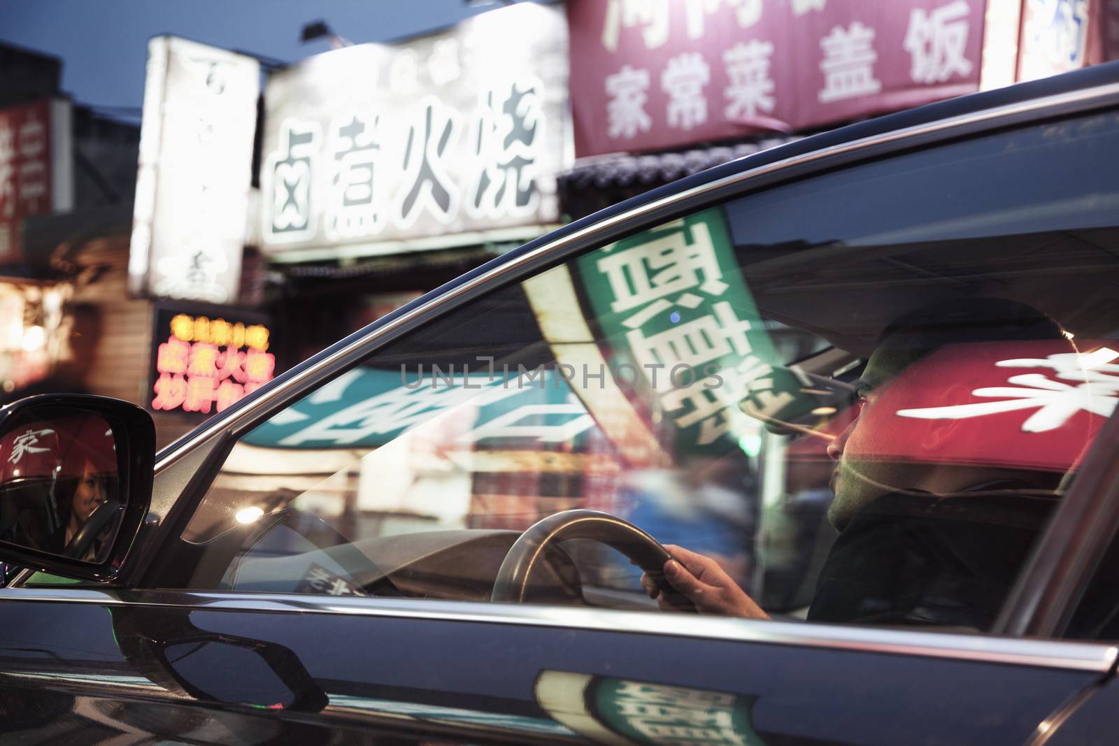 Young man driving through Beijing at night, illuminated store signs reflected off the windows of the car by XiXinXing