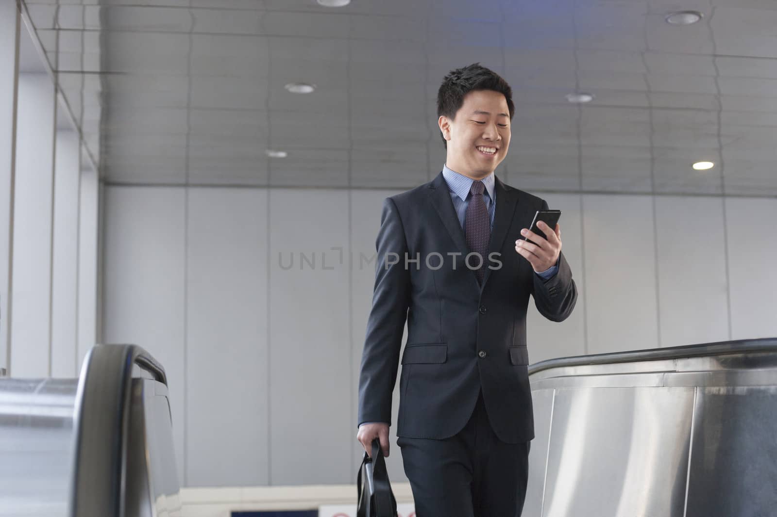 Smiling businessman coming up the escalator and looking down at his phone