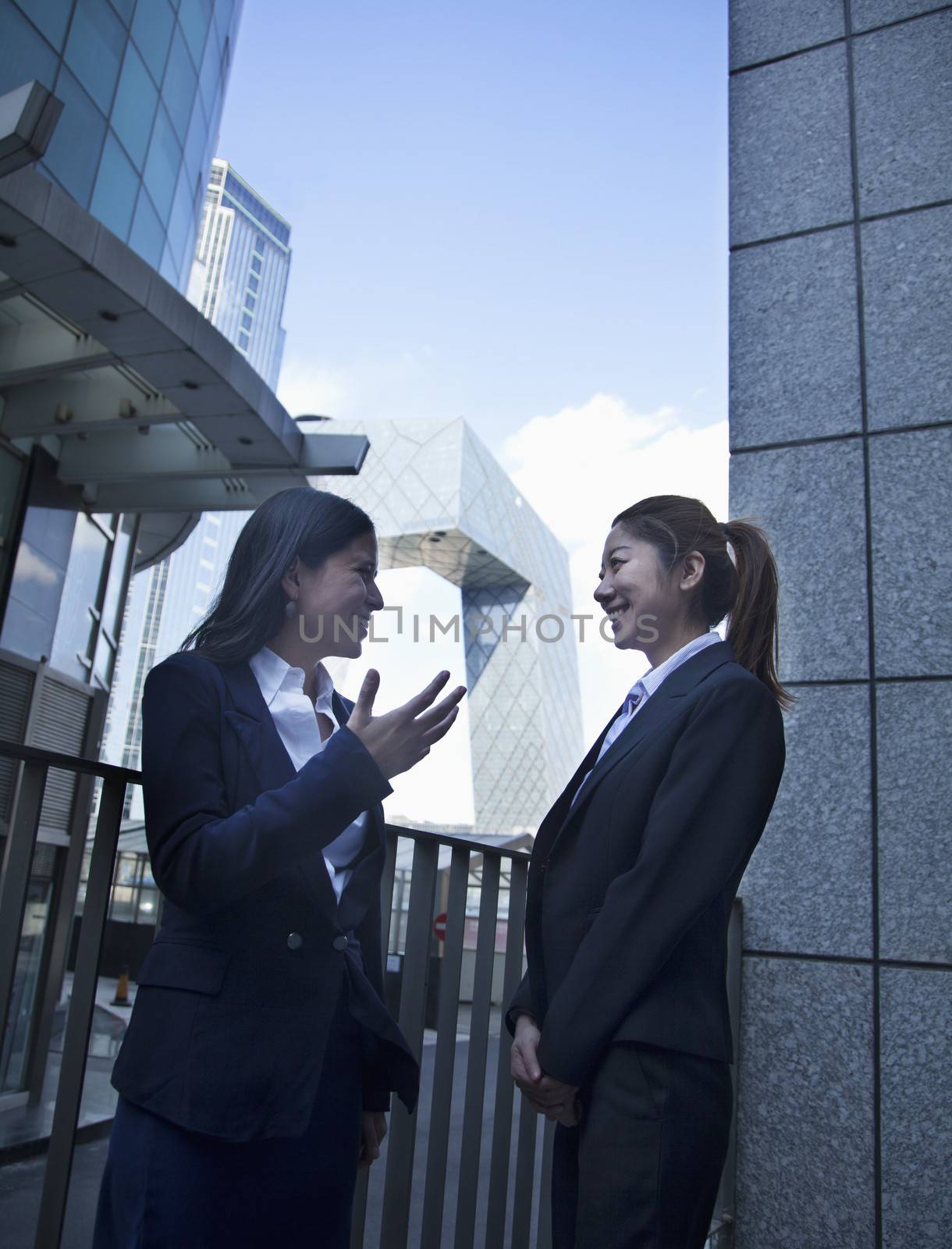 Two young confident businesswomen talking outdoors in Beijing, with CCTV building in the background