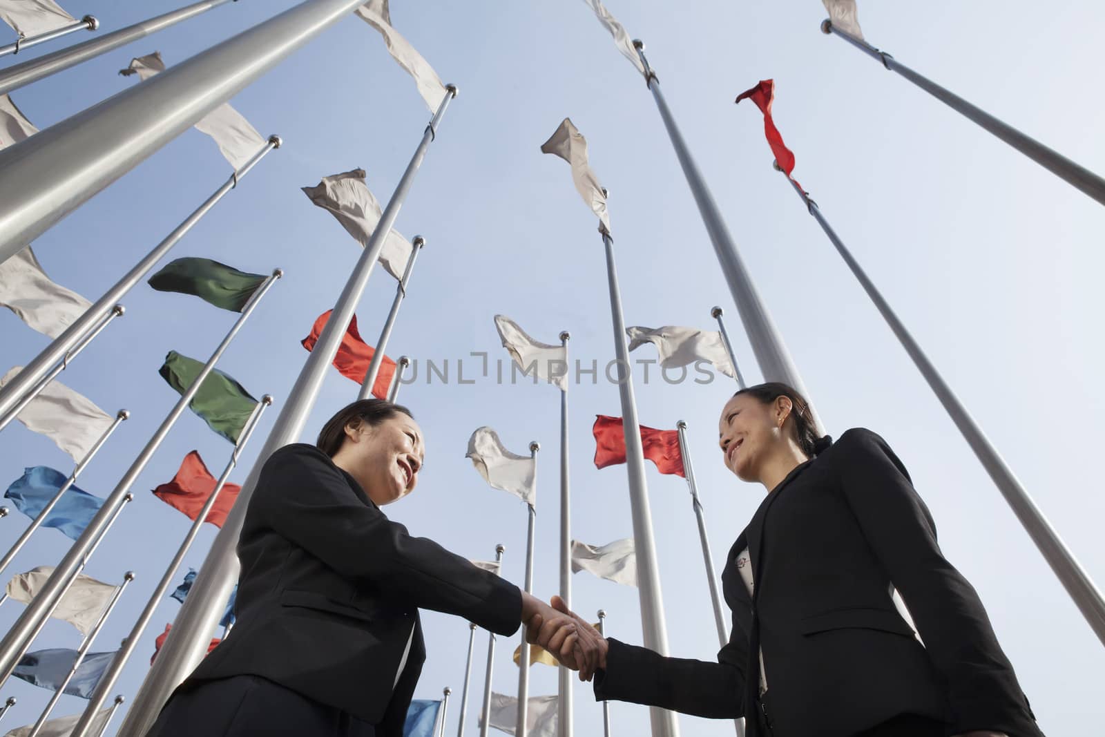 Two businesswomen shaking hands with flags in background. by XiXinXing