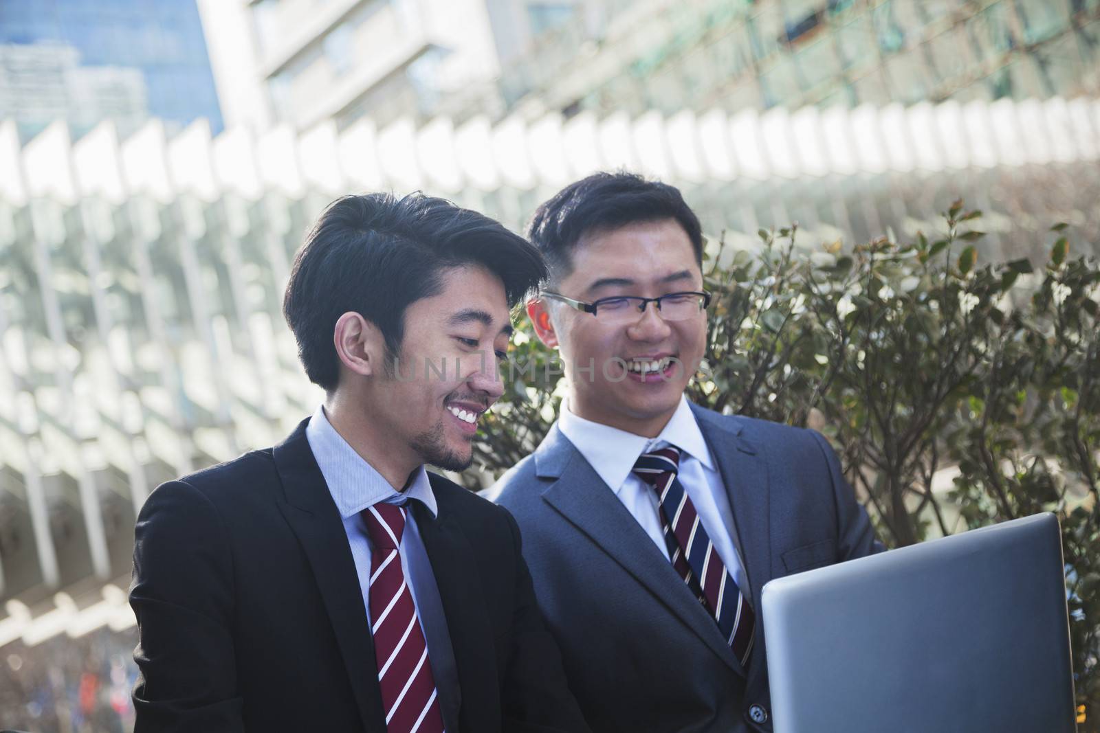 Two smiling businessmen looking at laptop together outdoors in Beijing by XiXinXing
