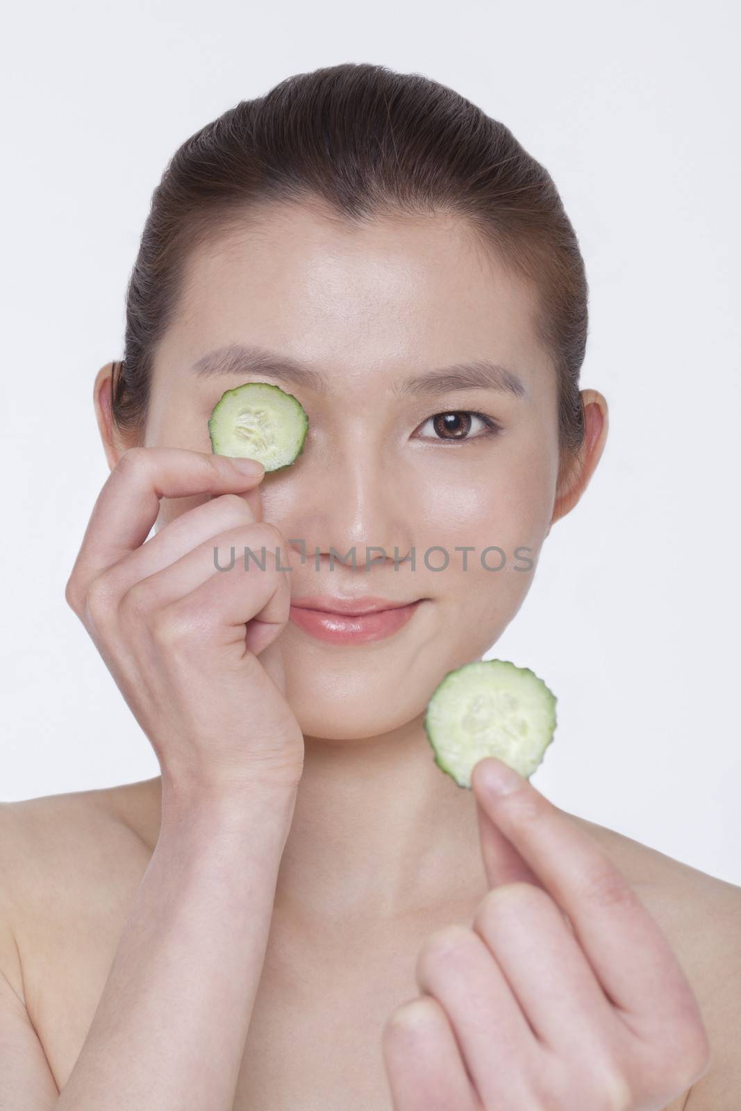 Smiling young woman holding up cucumber slices to put over her eyes, studio shot by XiXinXing