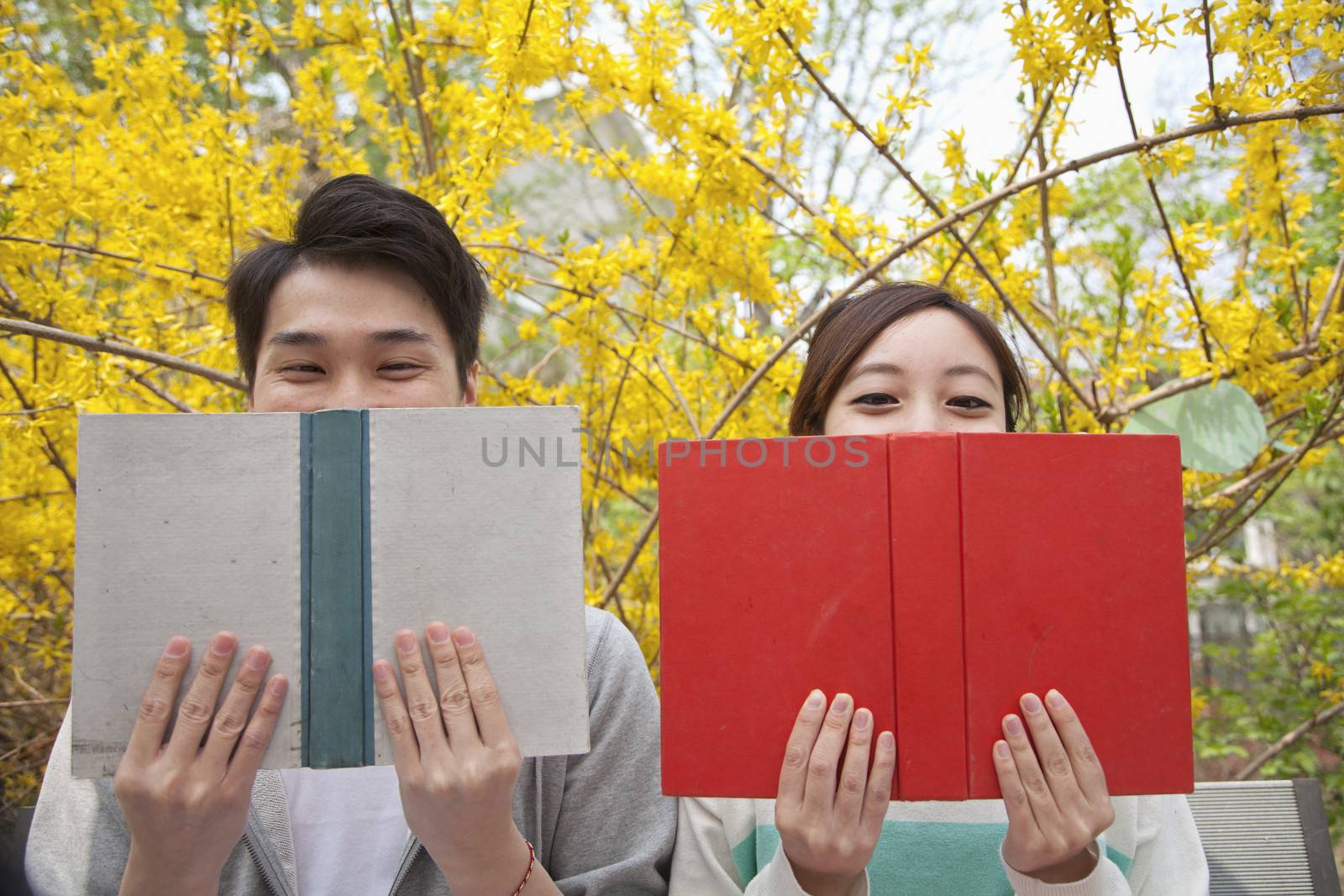 Young happy couple sitting side by side and reading their books, face partially obstructed, outdoors in springtime by XiXinXing
