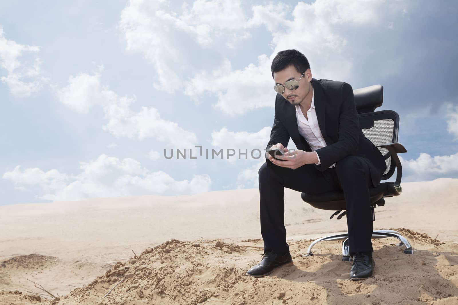 Young businessman in sunglasses sitting in an office chair in the middle of the desert, checking his phone