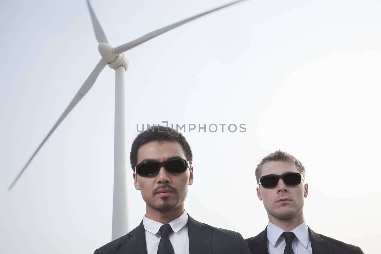 Portrait of two serious young businessmen in sunglasses standing by a wind turbine by XiXinXing