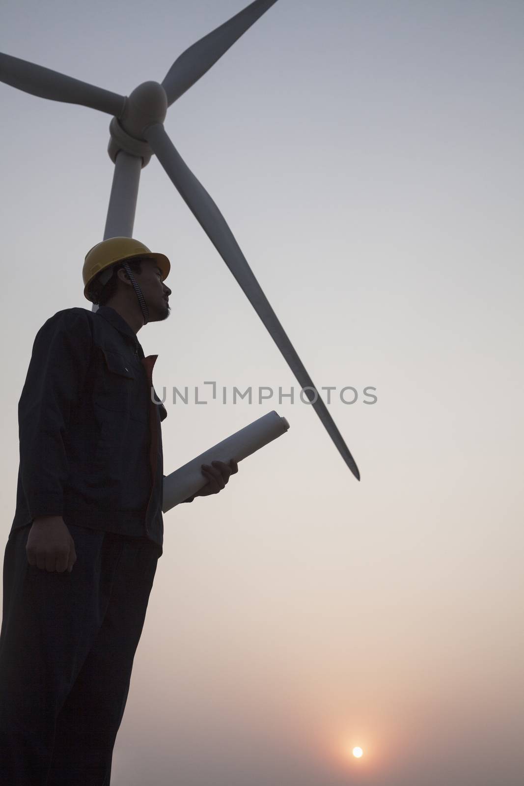 Silhouette of young male engineer holding a blueprint and standing beside a wind turbine at sunset by XiXinXing