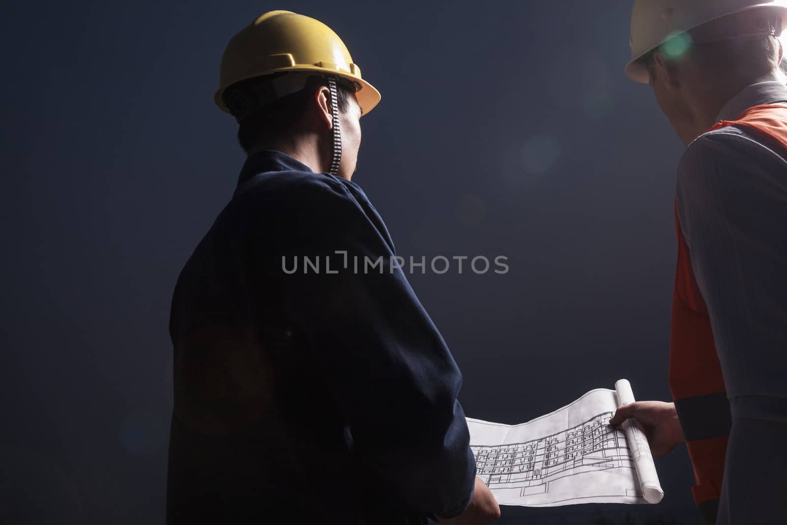 Two young male engineers in hardhats looking down at a blueprint at dusk, rear view by XiXinXing