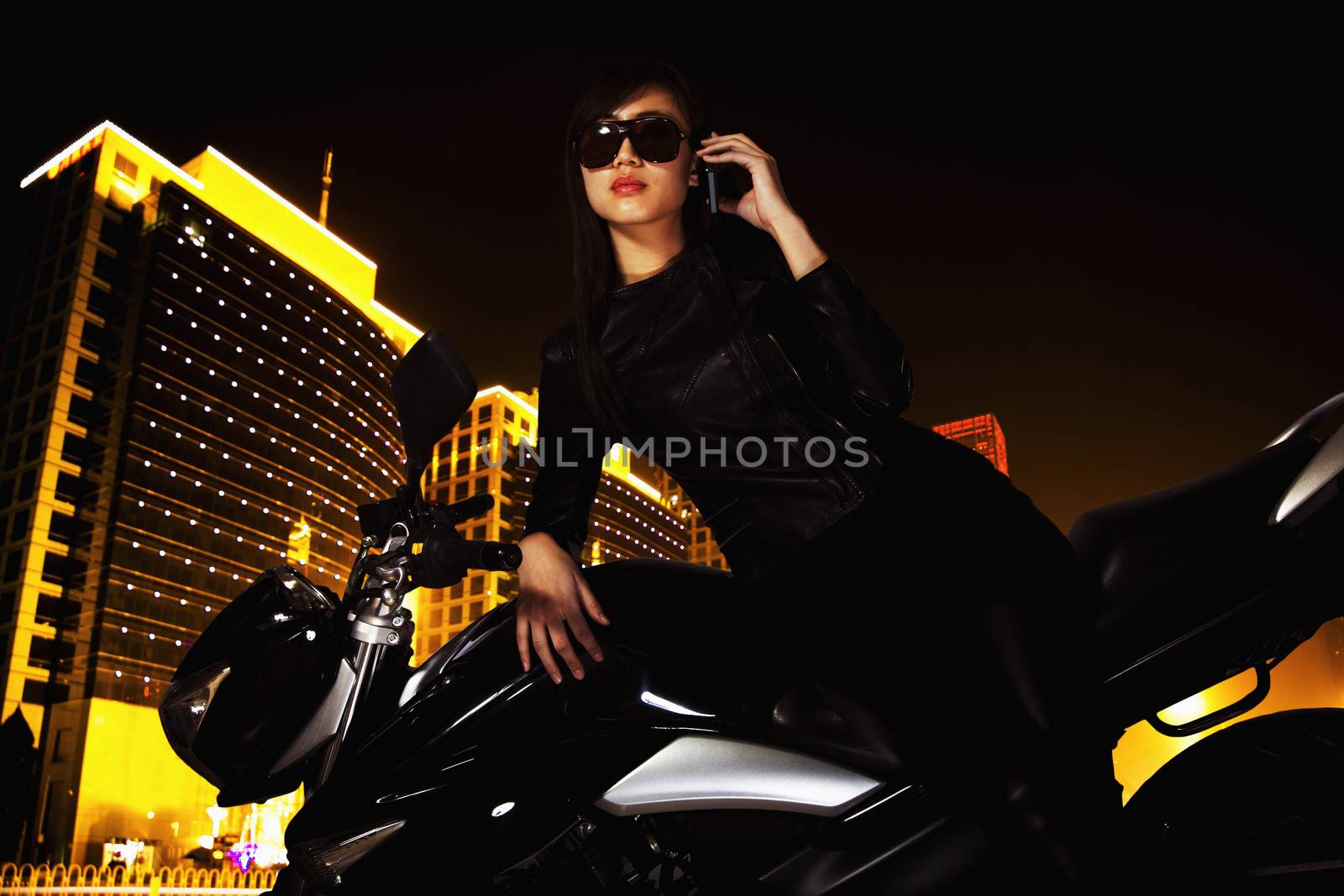 Beautiful young woman with sunglasses talking on the phone and leaning on her motorcycle at night