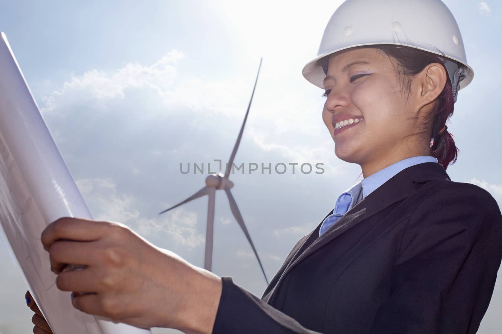 Young smiling female engineer holding open and looking down at blueprints, on site with wind turbines by XiXinXing