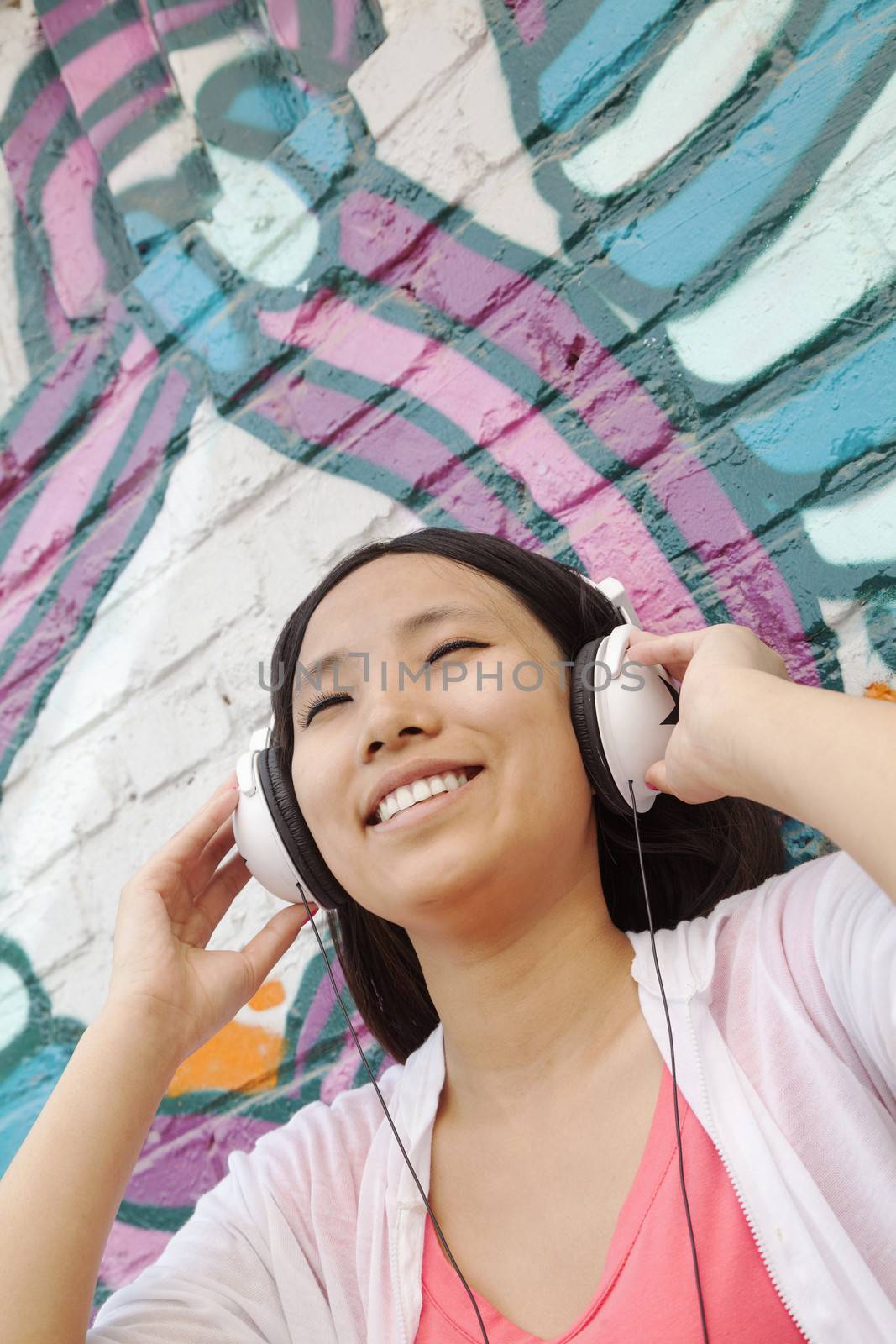 Young smiling woman holding her headphones while enjoying listening to music in front of wall with graffiti by XiXinXing