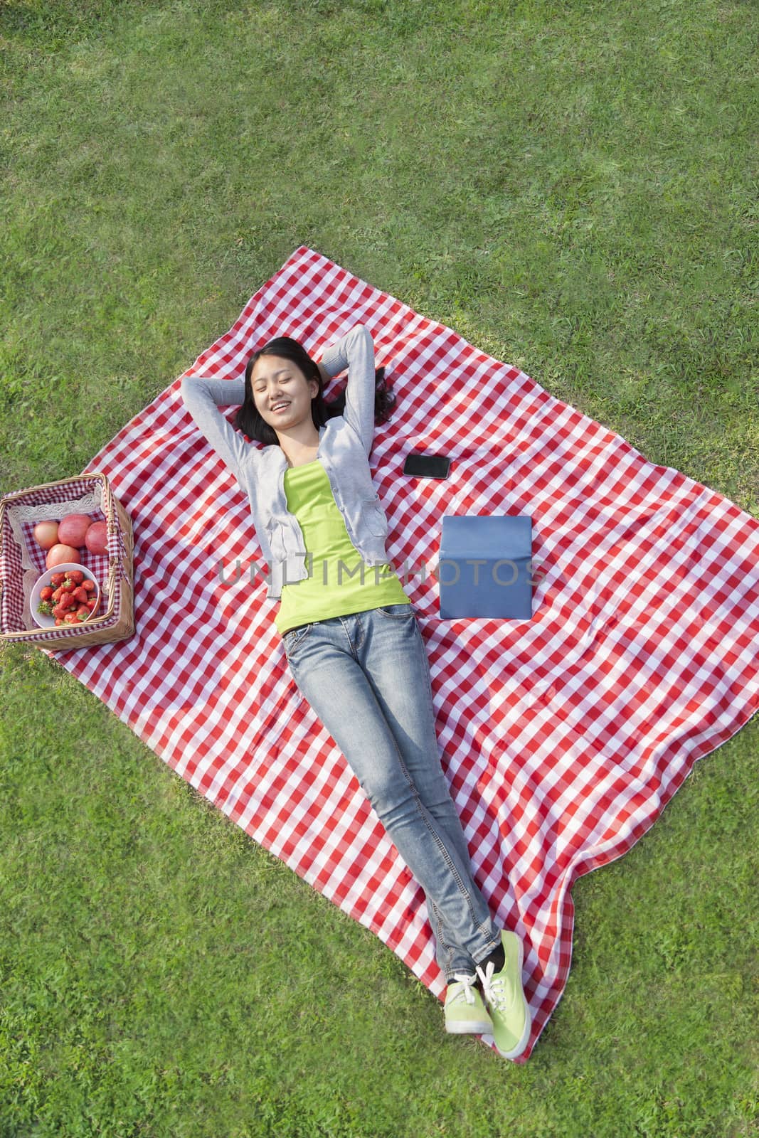 Smiling young woman lying on her back with arms behind her head on a blanket and relaxing in the park