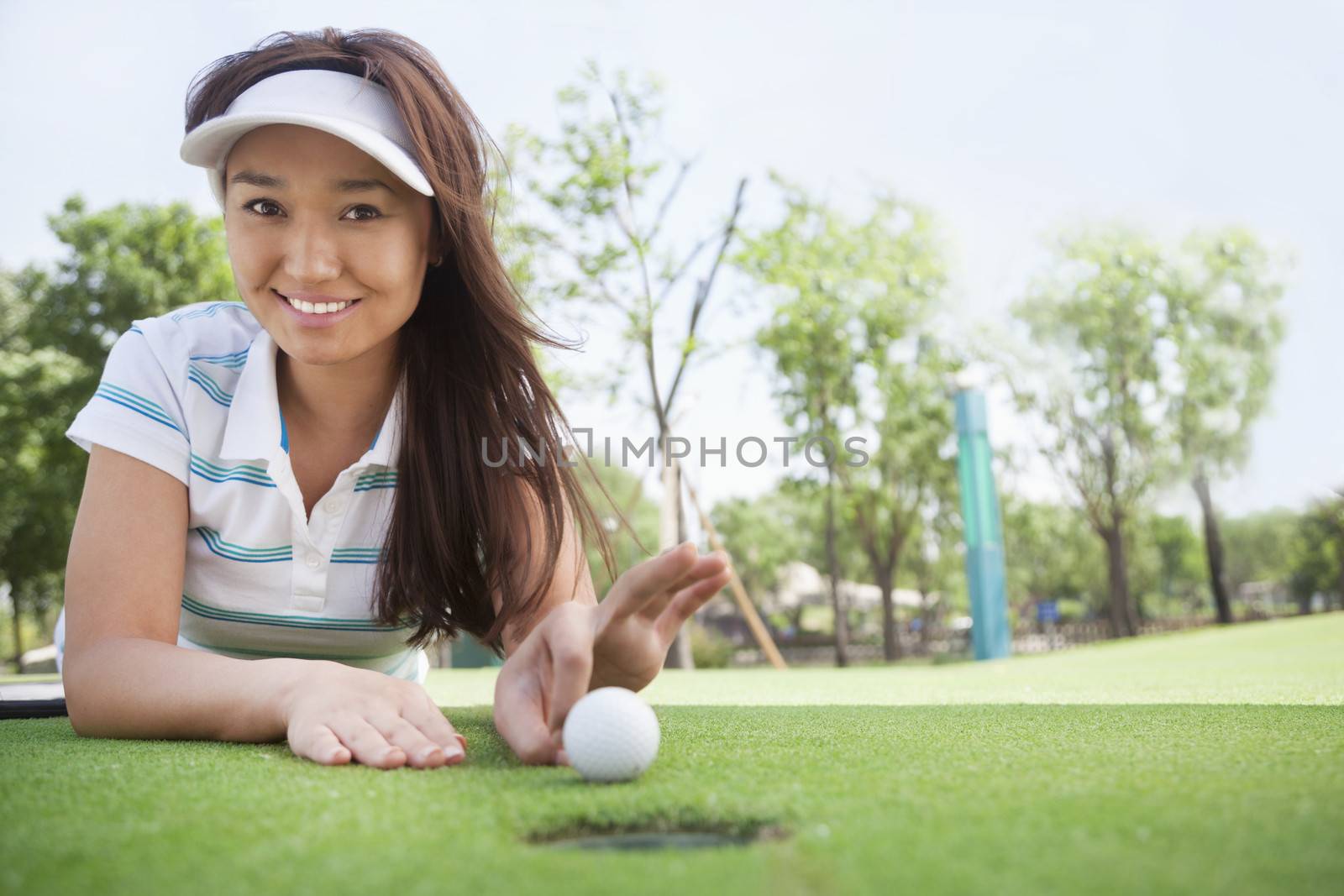 Smiling young woman lying down in a golf course getting ready to flick the ball into the hole