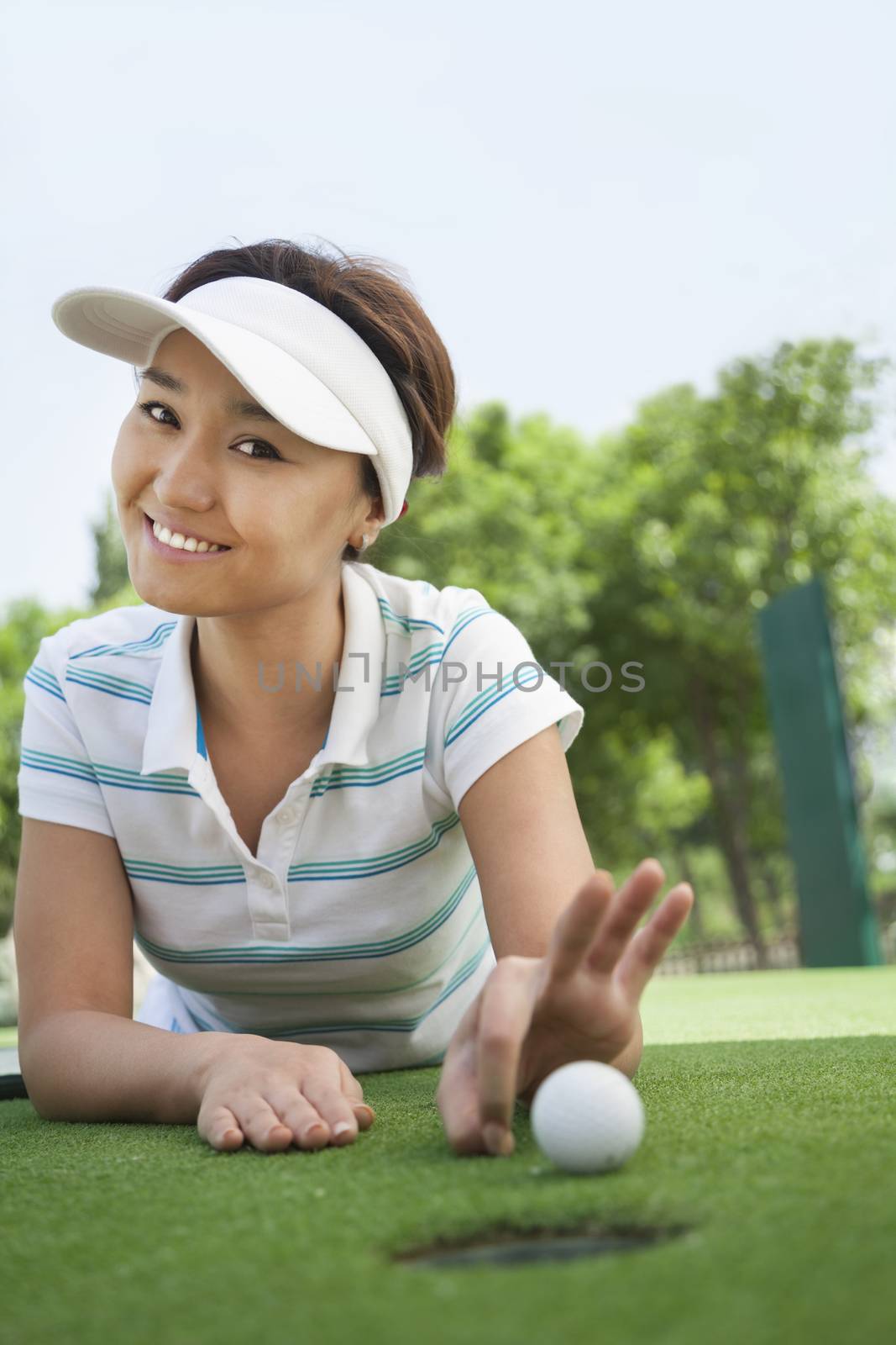 Smiling young woman lying down in a golf course getting ready to flick the ball into the hole by XiXinXing
