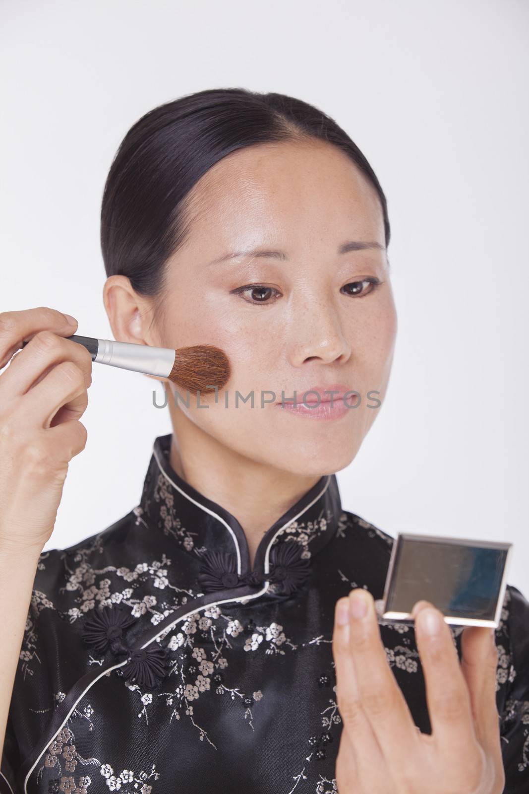 Woman in traditional clothing looking into a mirror and applying make up with a make up brush, studio shot